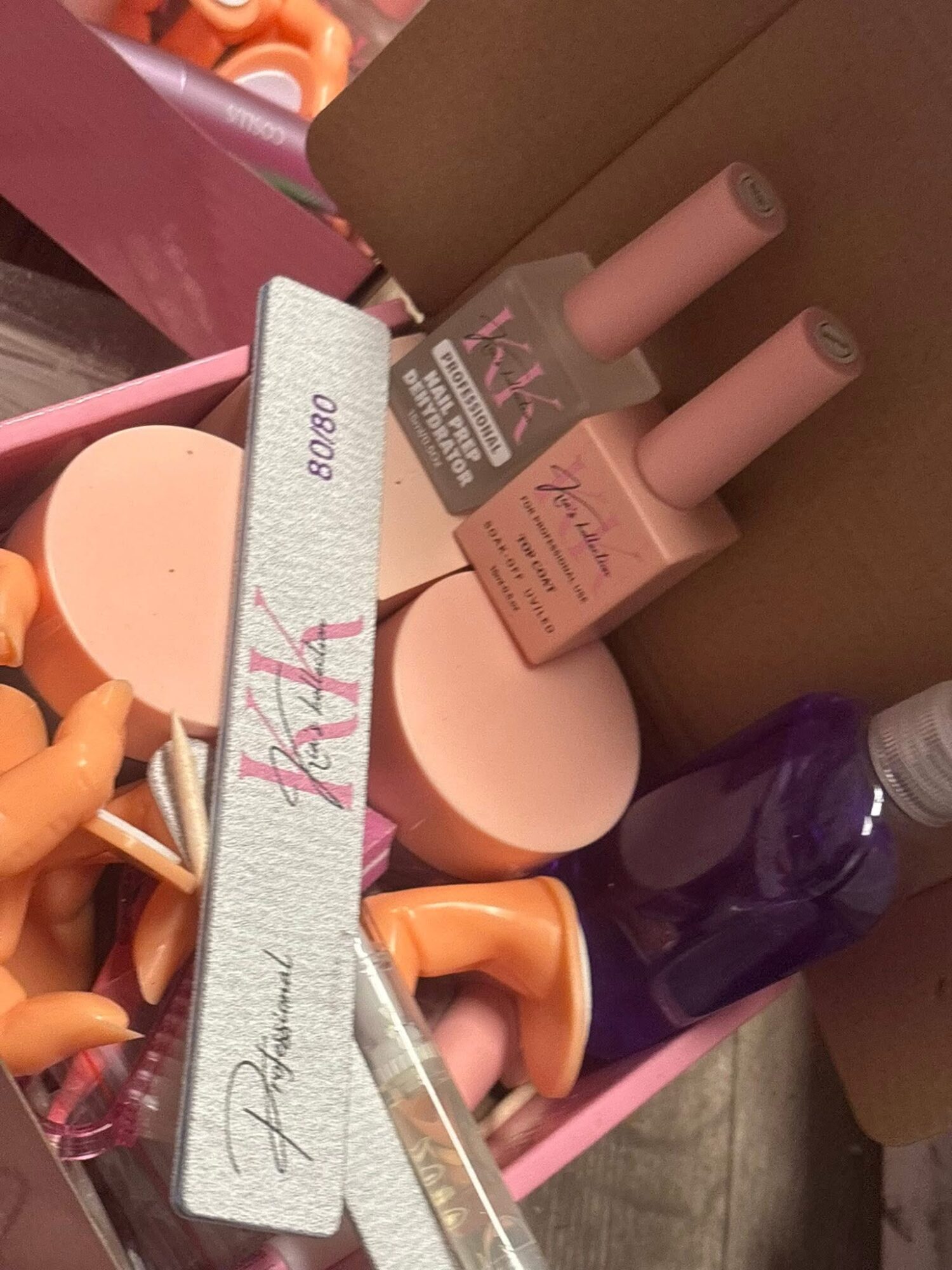 Open pink box containing nail polish bottles, a round container, and orange objects, with a purple bottle visible.