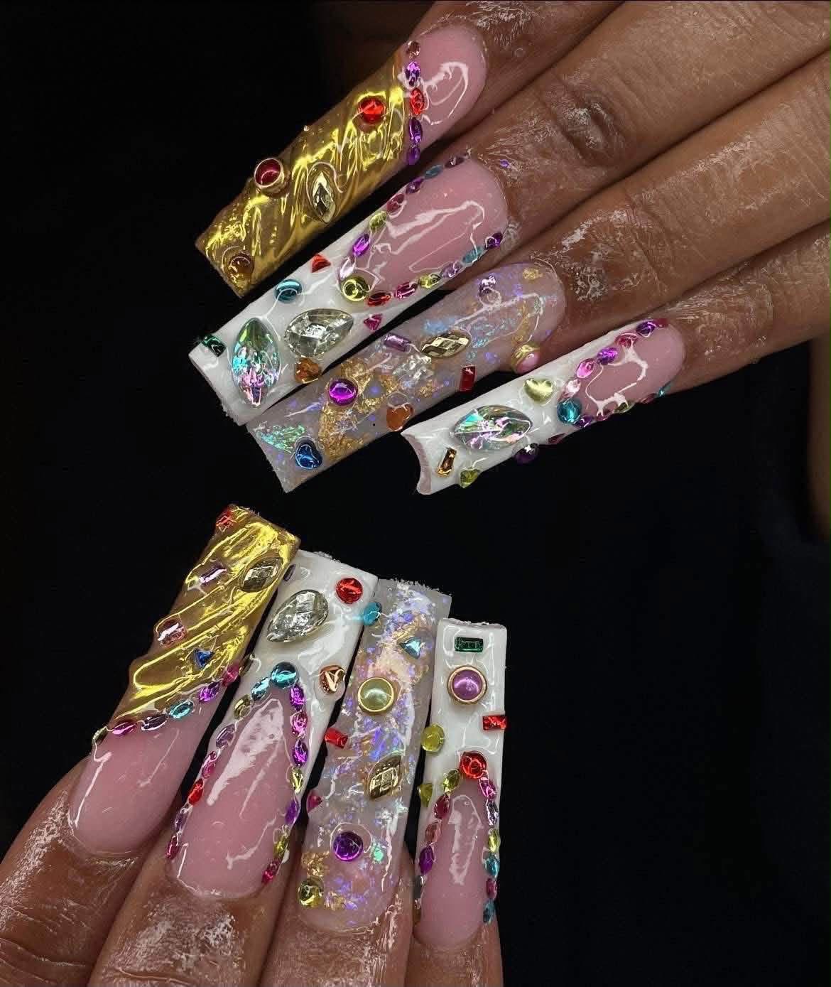 Close-up of decorated long nails with gold, silver, and colorful rhinestones on a person's hand, against a black background.