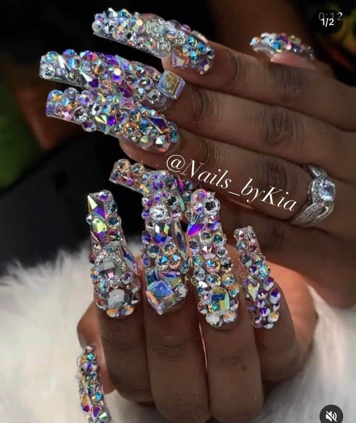 Close-up of hands with long, decorated nails covered in multicolored rhinestones and jewelry, with a ring on one finger.