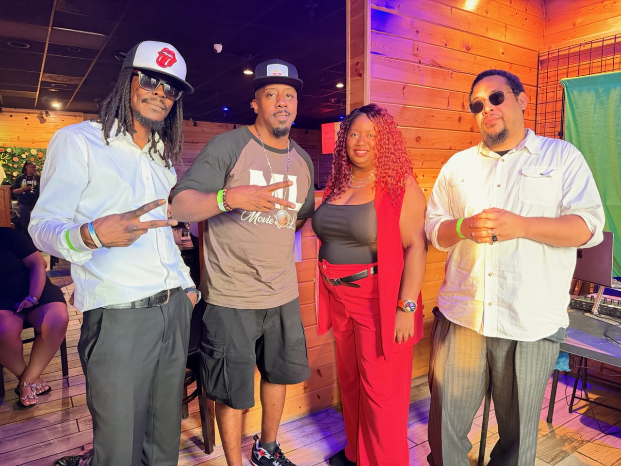 Four people standing indoors, two men and two women, with wooden walls and colorful lighting in the background.