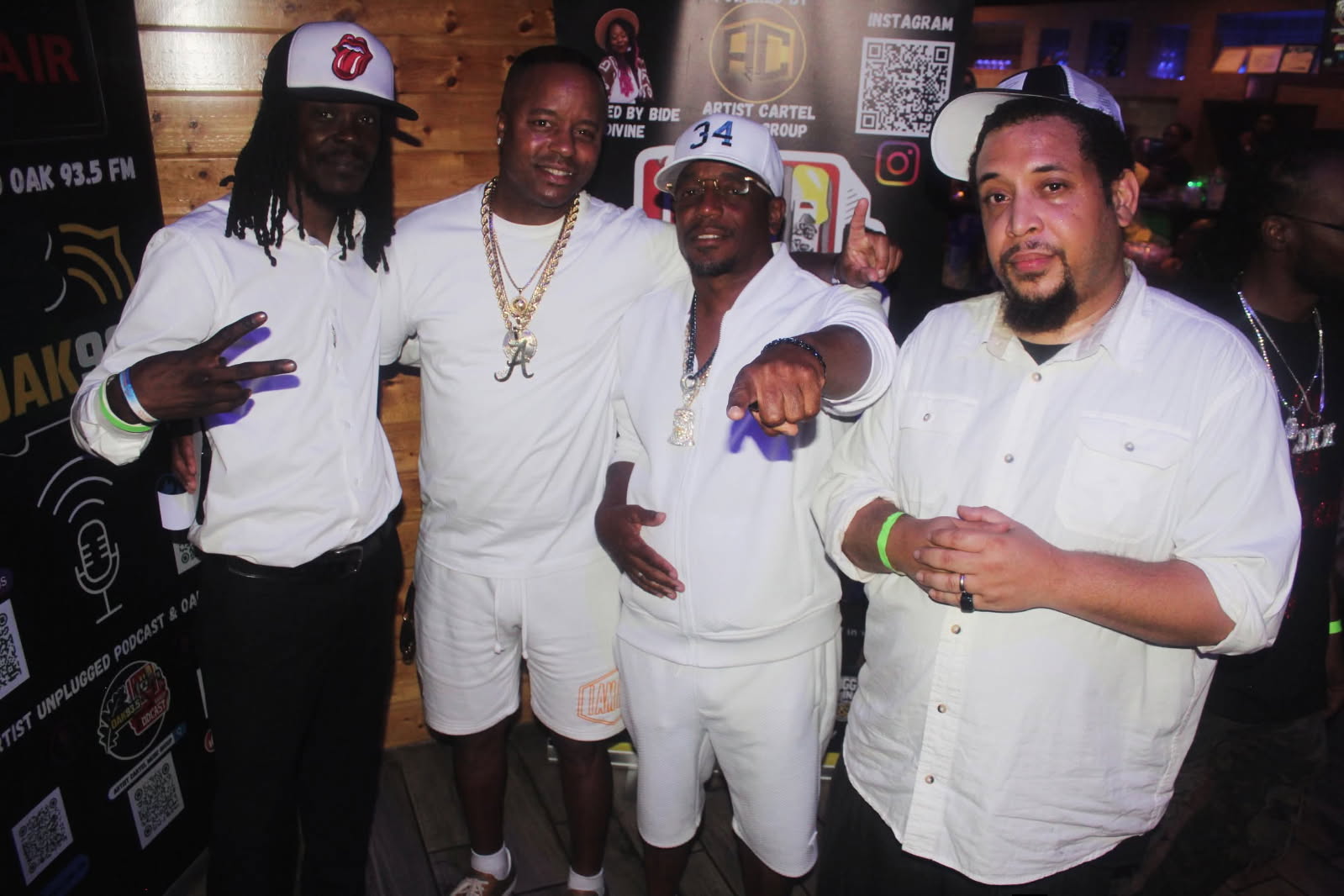 Four men in white clothing and hats posing together indoors, some making hand gestures, smiling at the camera.