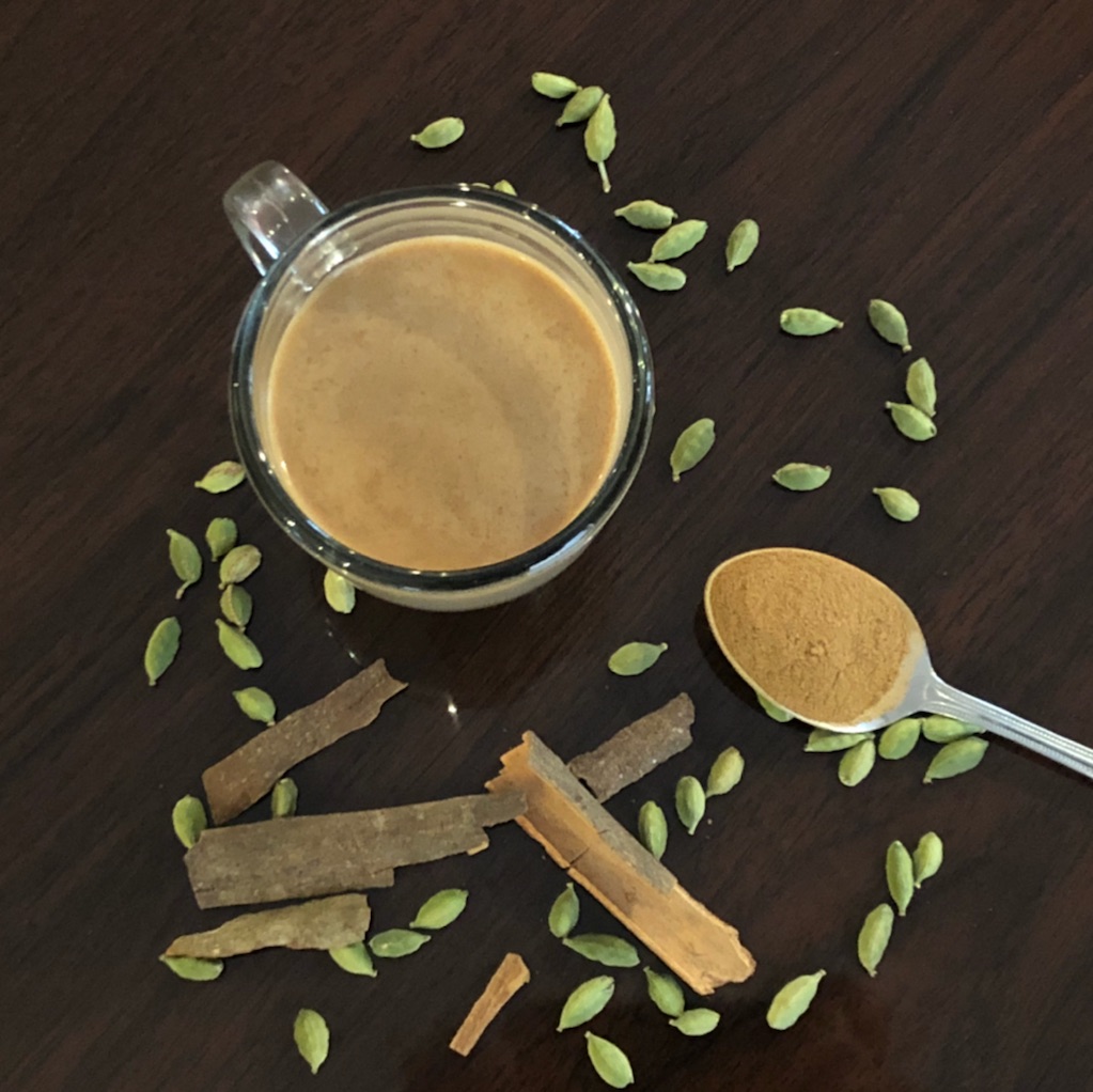 A cup of coffee, cinnamon sticks, green cardamom pods, and a spoonful of ground cinnamon on a dark wooden surface.