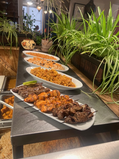 Three white plates with grilled meat, spicy chicken, and side dishes on a black table, with green plants in the background.