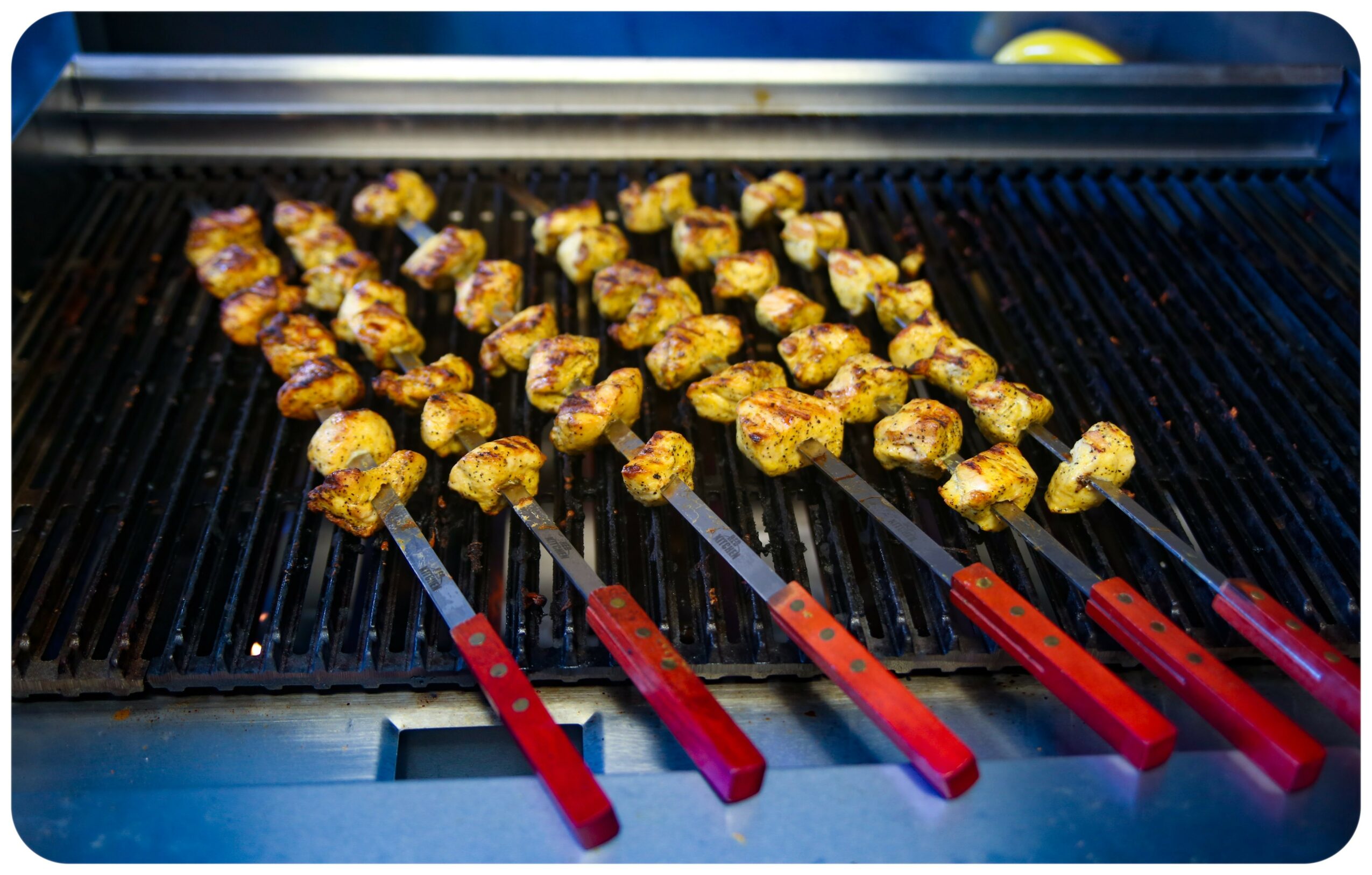 Skewers with grilled meat cooking on a barbecue grill, with a black grate and red handles.