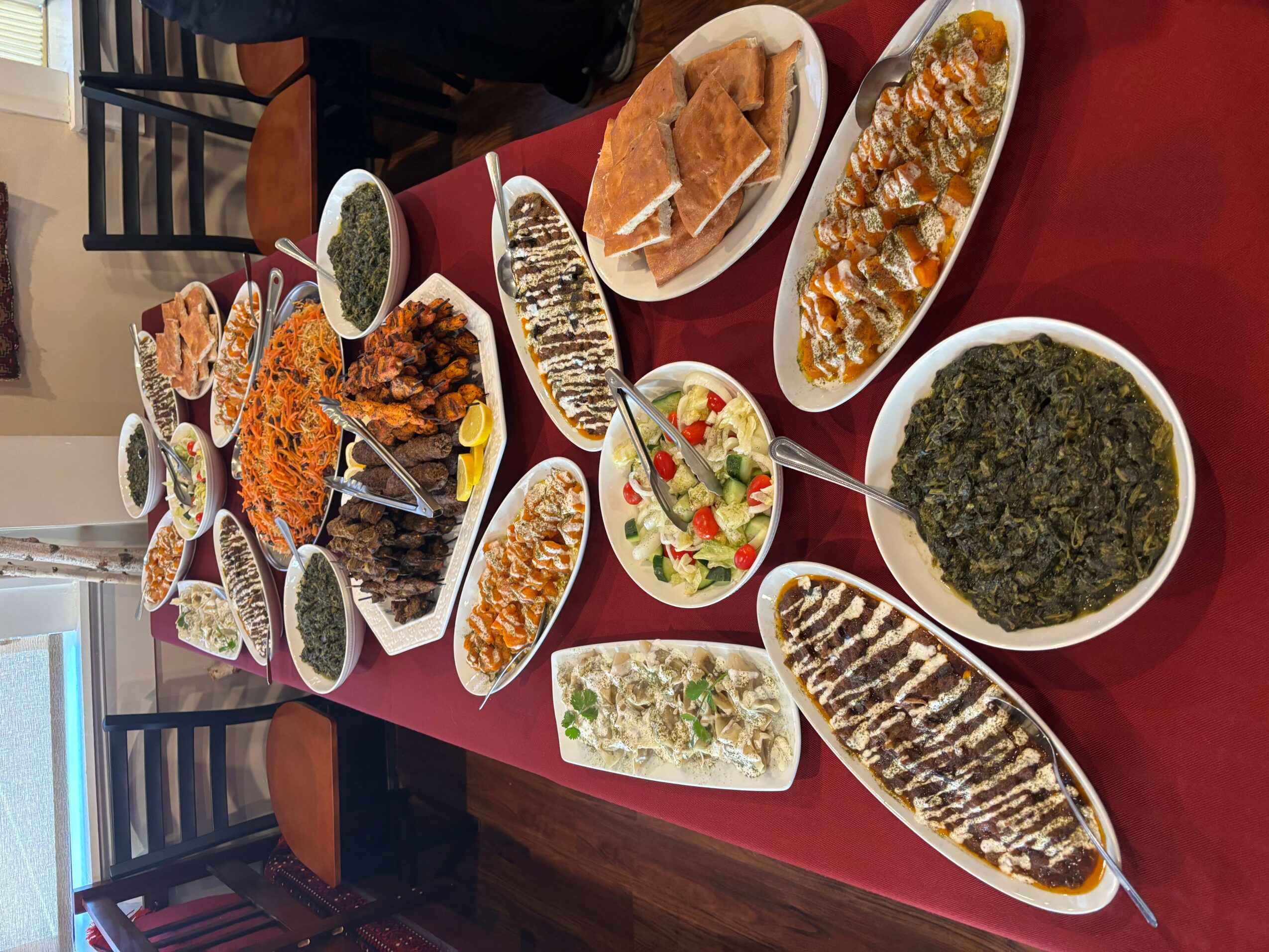 Assorted dishes including grilled fish, salads, and cooked greens on a red table, with chairs in the background.