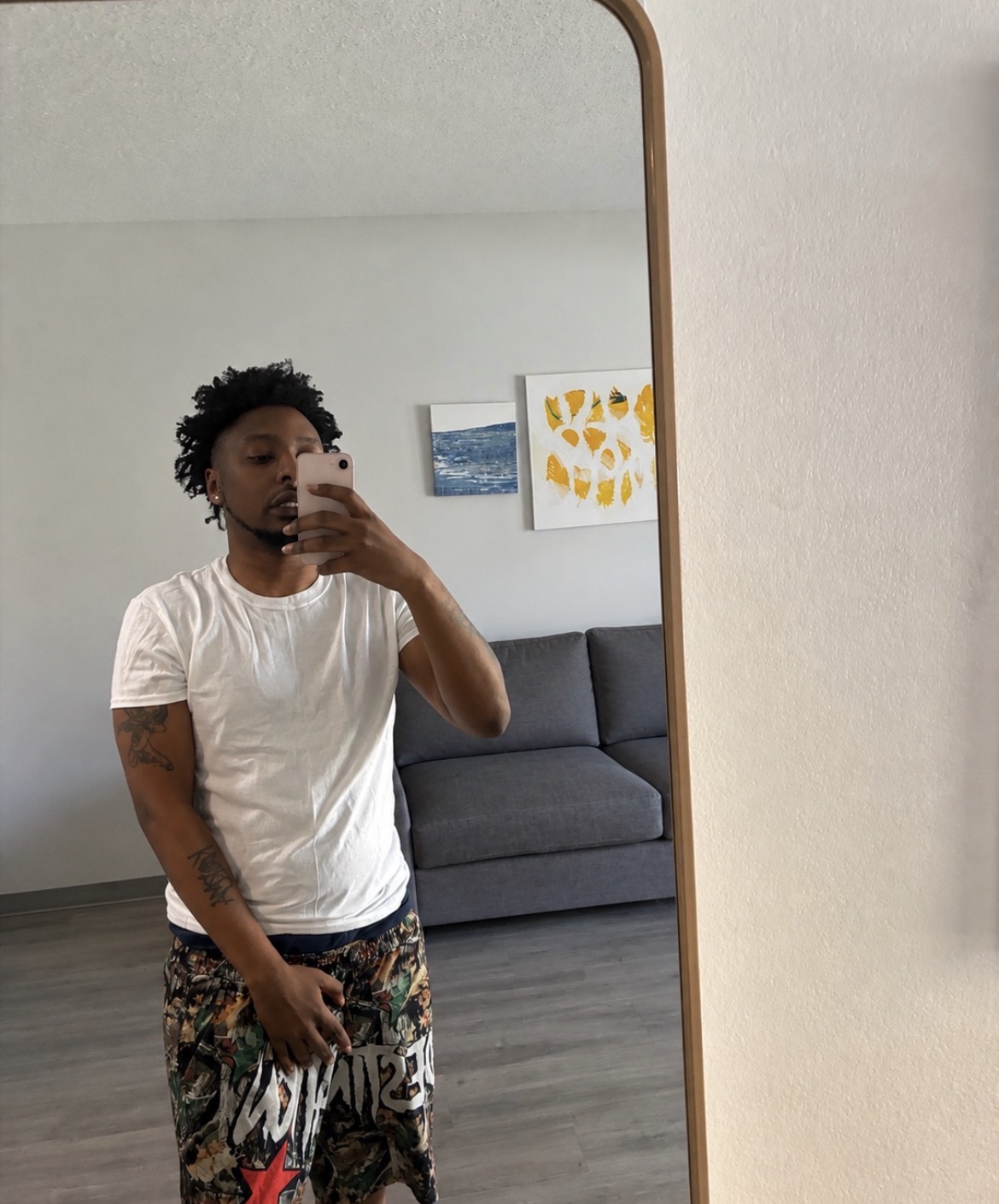 Person with short curly hair taking a mirror selfie in a room with a gray sofa and artwork on the wall.