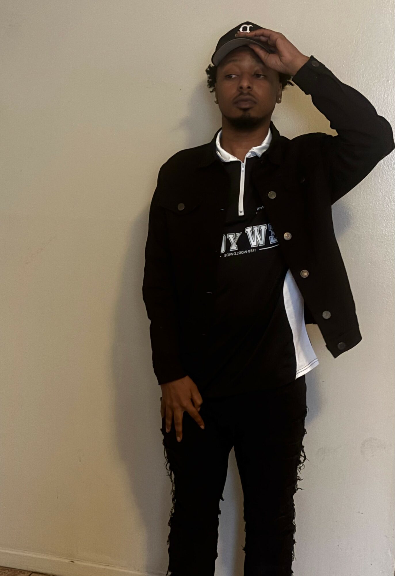 Young man standing against a plain wall, wearing a black jacket, black pants, and a cap, with one hand on his head.