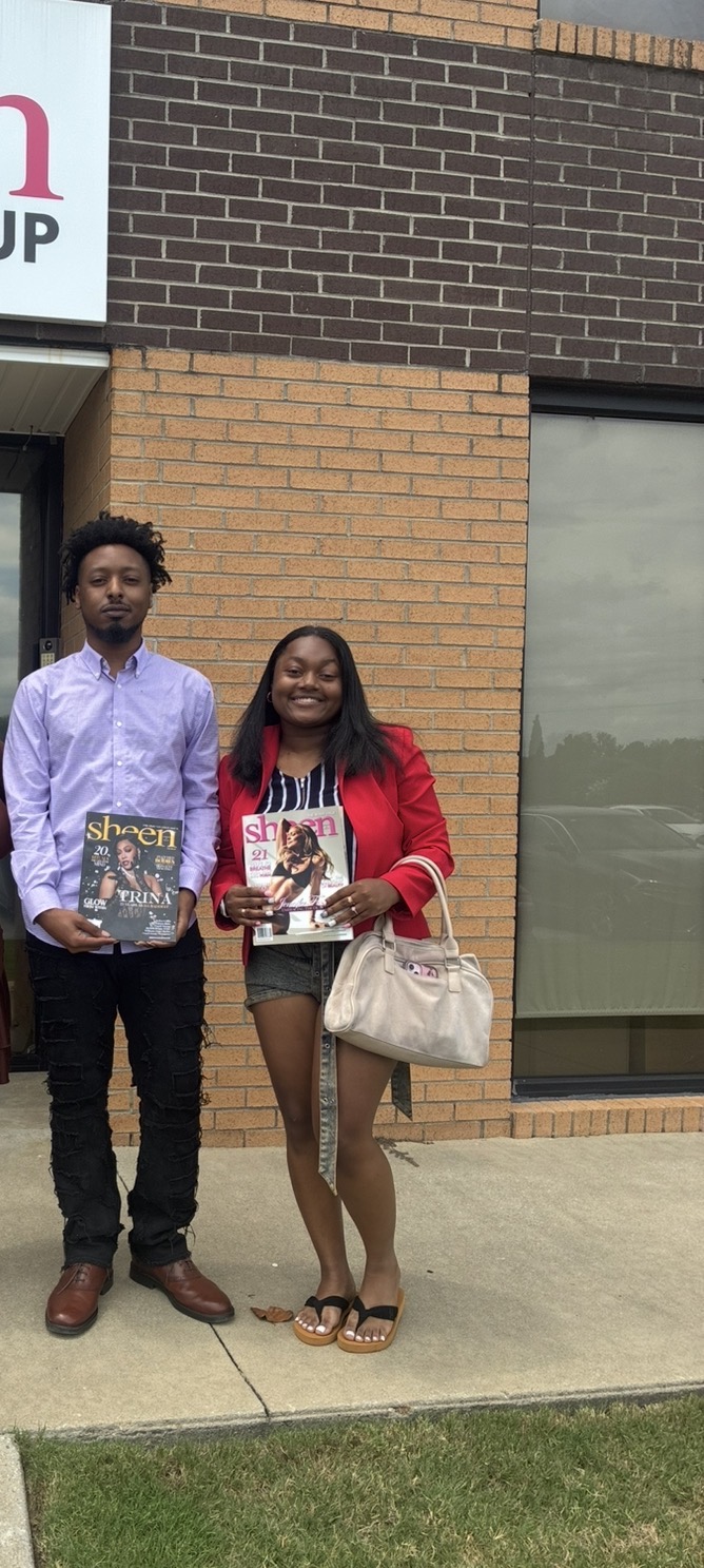 Two people stand outside a building holding magazines, with a brick wall and window behind them.