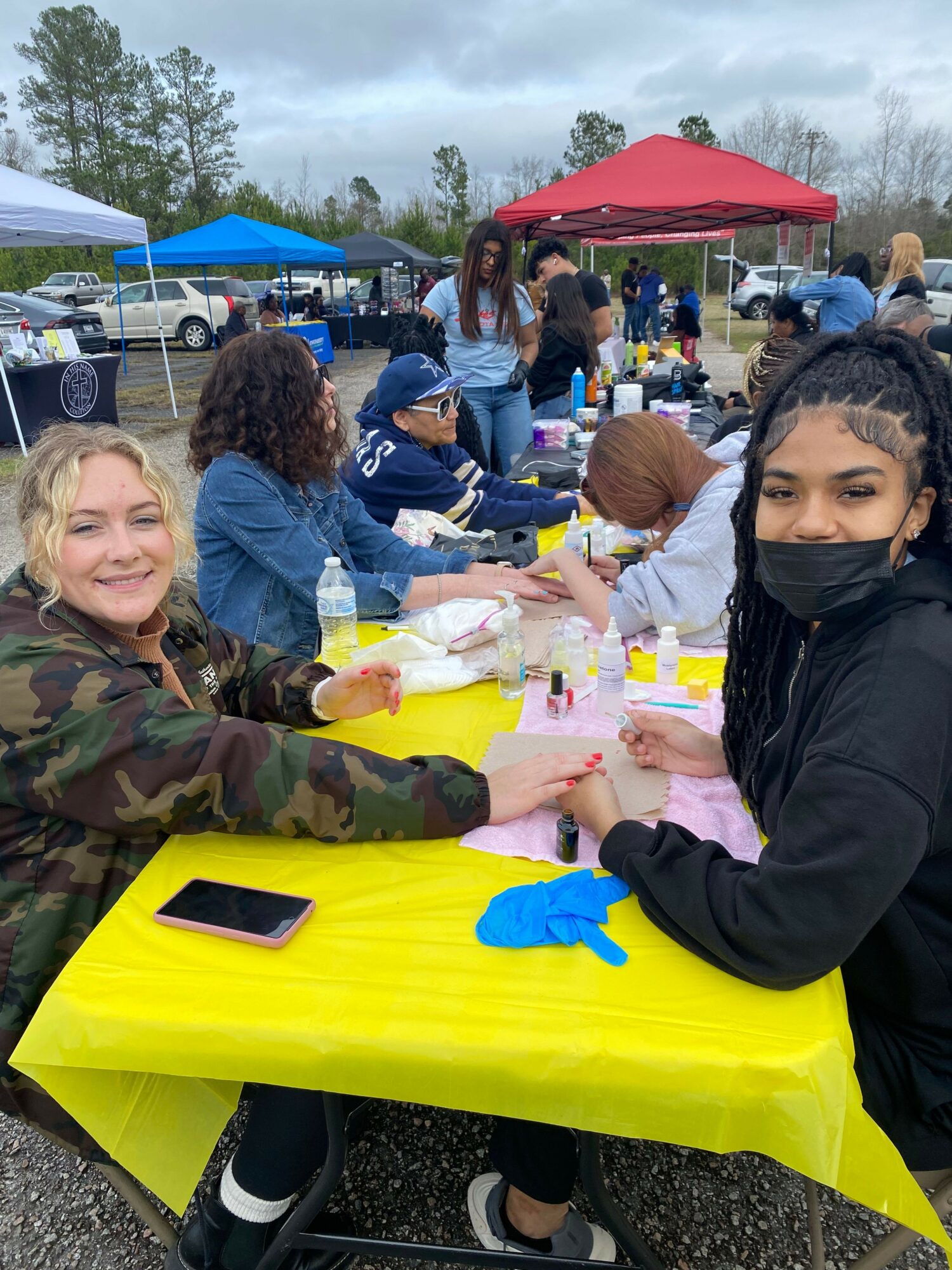 Two women sitting at a yellow table outdoors, one wearing a black mask and the other in camouflage, with people and tents in background.