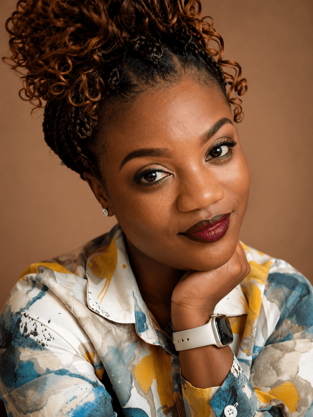 Portrait of a woman with curly hair, wearing a colorful shirt, resting her chin on her hand, against a brown background.