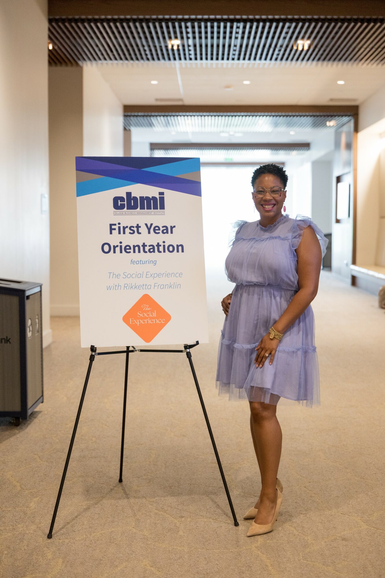 Woman in a purple dress standing next to a sign for first-year orientation, smiling, in a modern indoor setting.