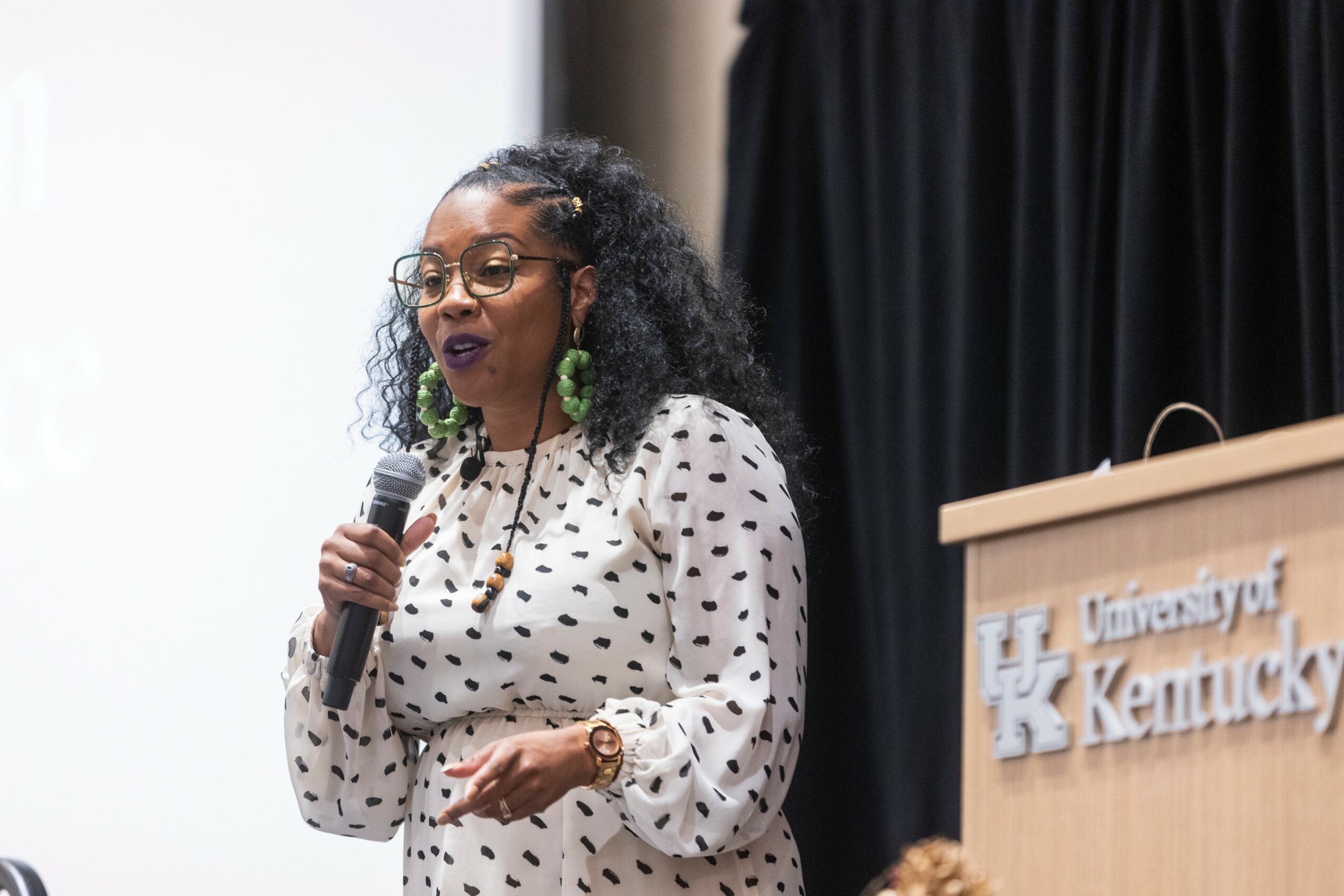 Woman speaking into a microphone, wearing glasses and a polka dot blouse, standing at a podium with University of Kentucky logo.