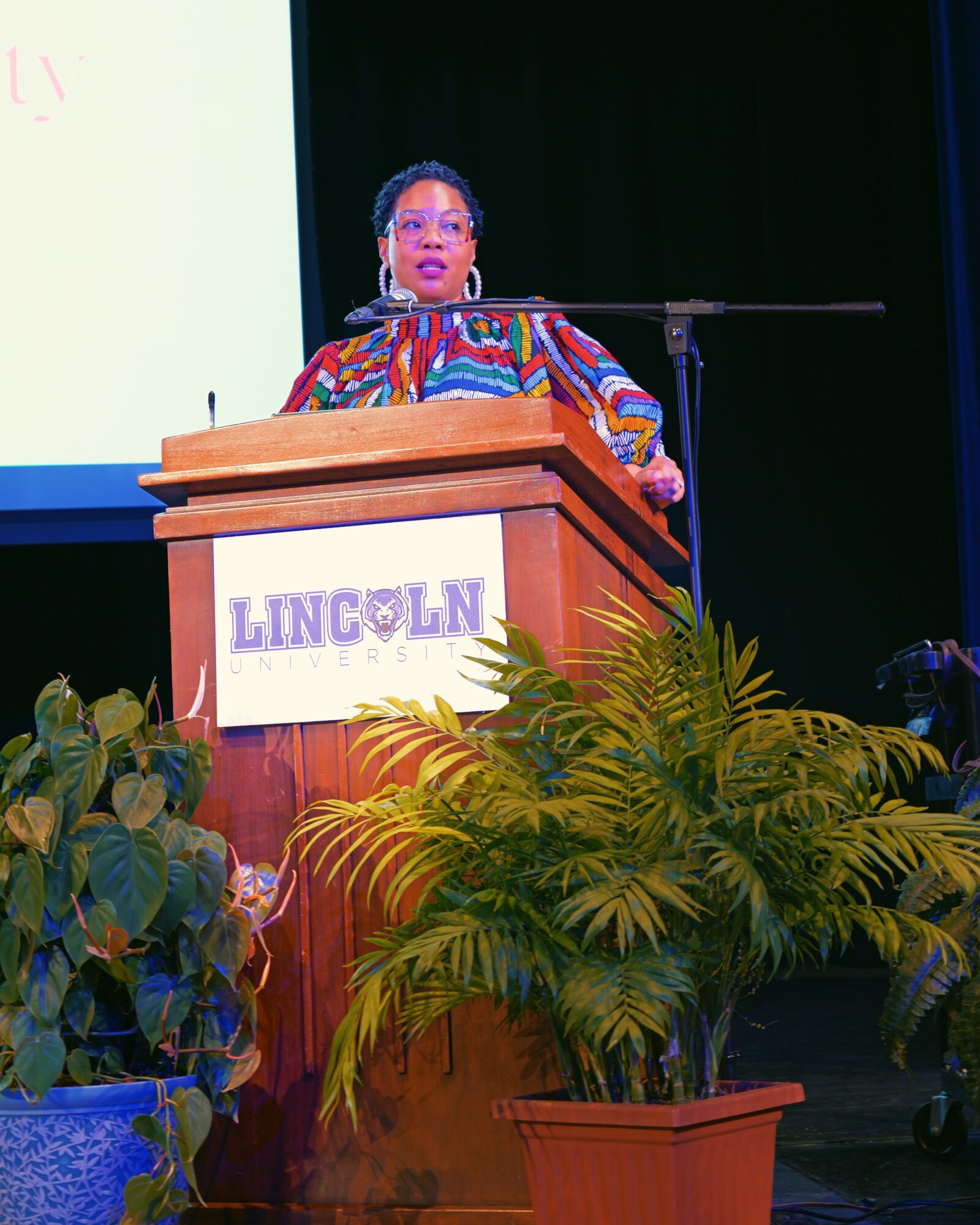 Woman speaking at a podium with a Lincoln sign, surrounded by green plants, on a stage with a black background.