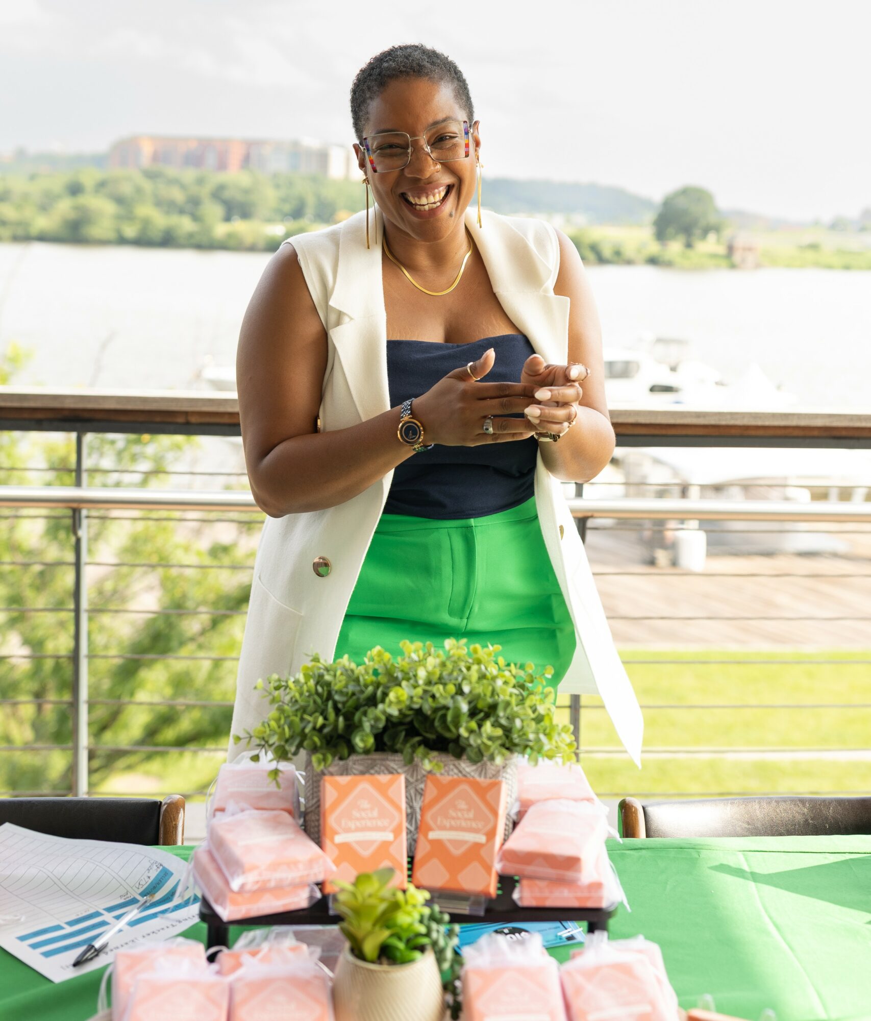 Smiling woman standing outdoors behind a table with gifts and plants, holding a phone, with a river and trees in background.