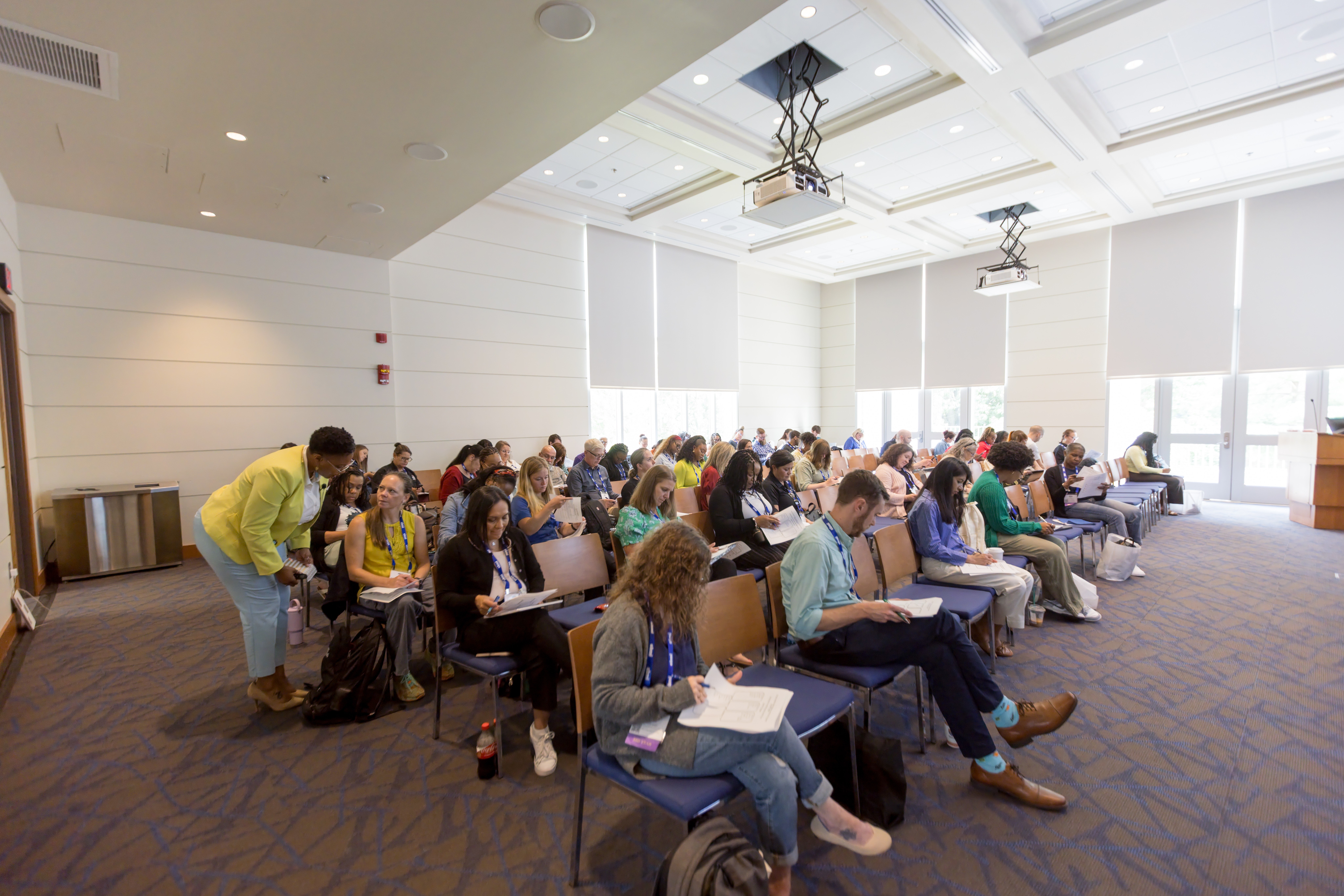Large classroom filled with students sitting at desks, taking notes, and listening attentively, with bright natural light from windows.