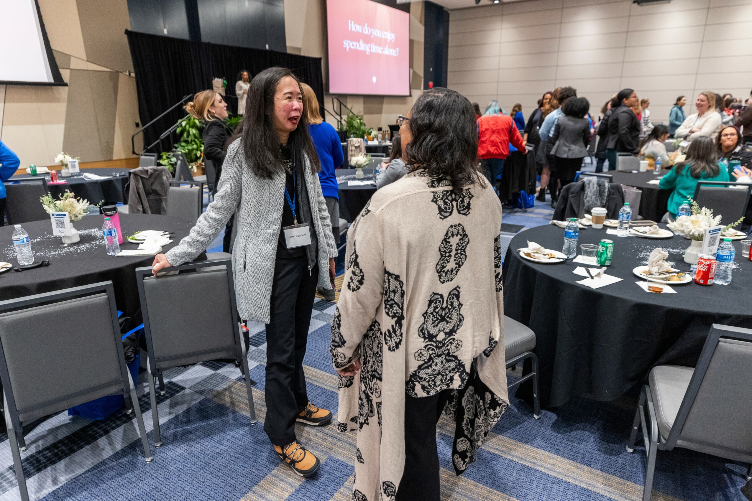 Two women are talking and smiling at a conference or event with round tables and many people in the background.