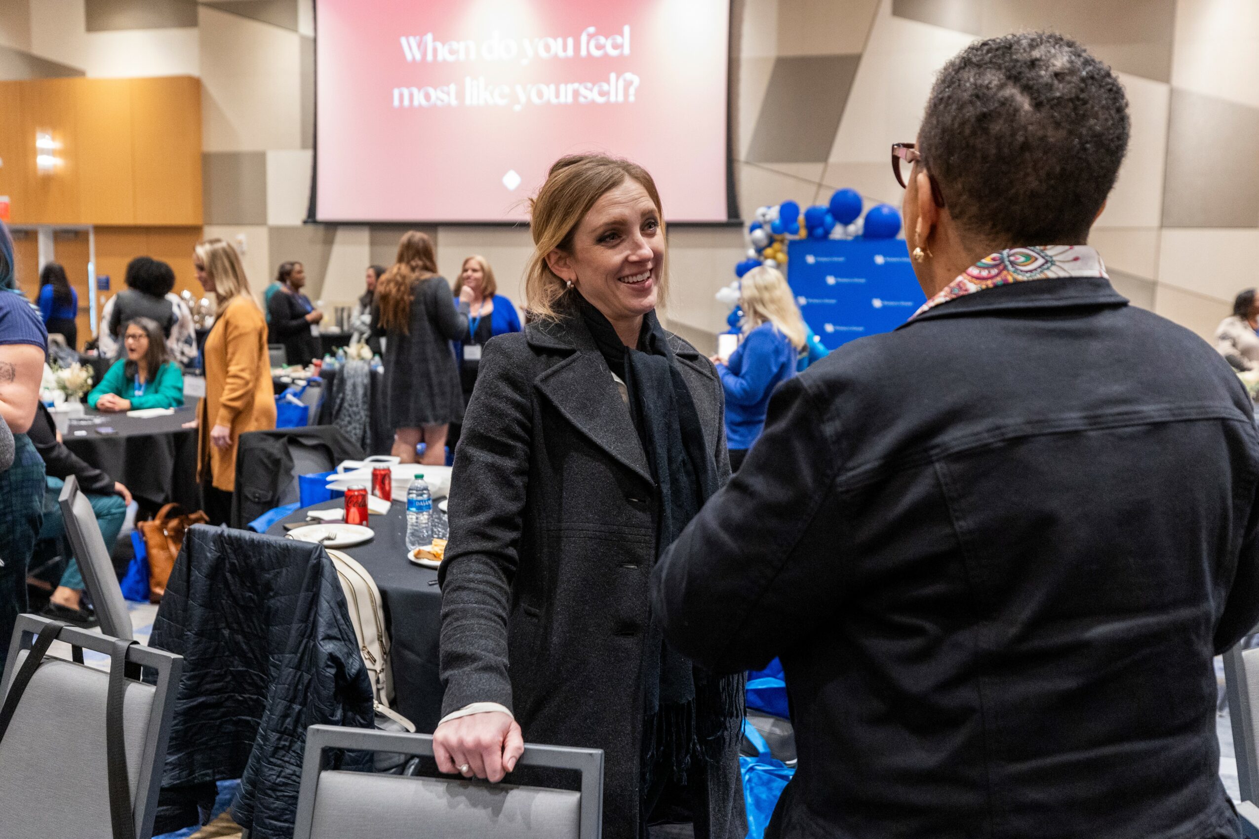 Woman smiling and talking to a man at a conference or event, with tables and a large screen in the background.