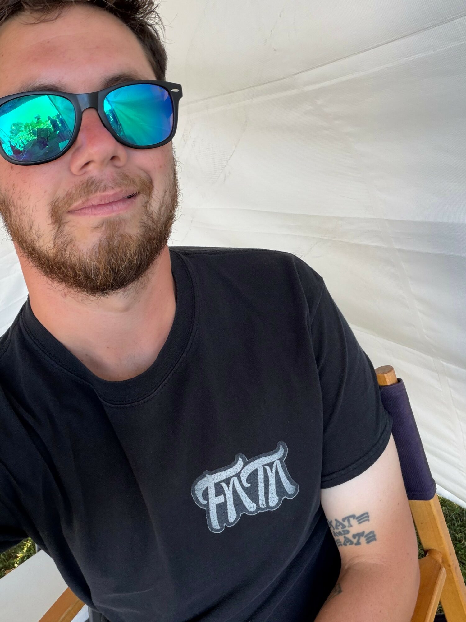 Young man with sunglasses and beard, wearing a black T-shirt, sitting outdoors with a white background.