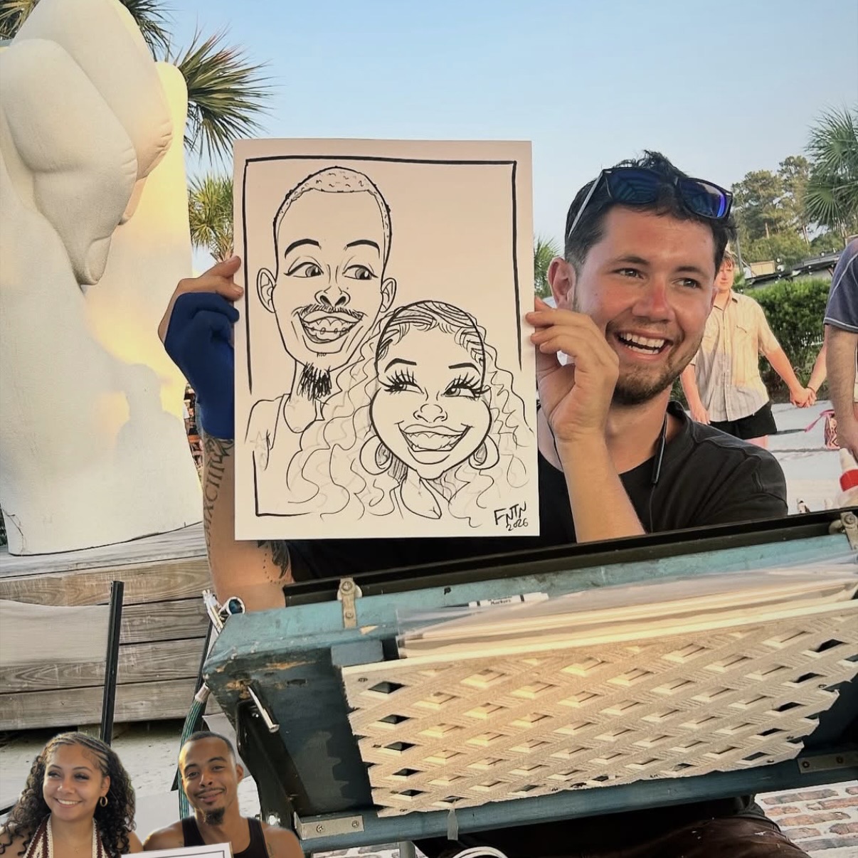 Person holding a caricature drawing of two smiling people outdoors, with palm trees and a sculpture in the background.