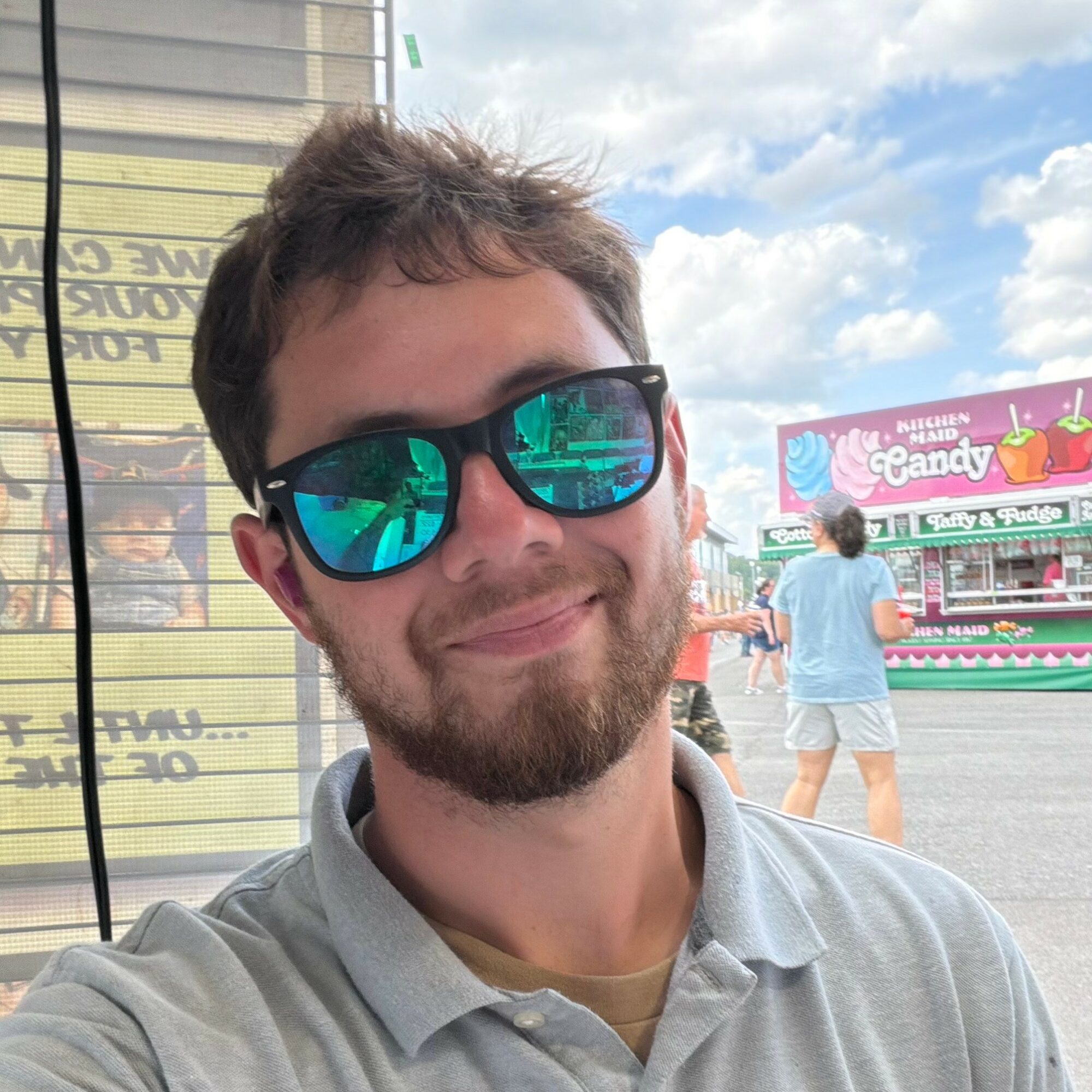 Young man with sunglasses smiling outdoors, background shows a fairground with a sign and a person walking away.