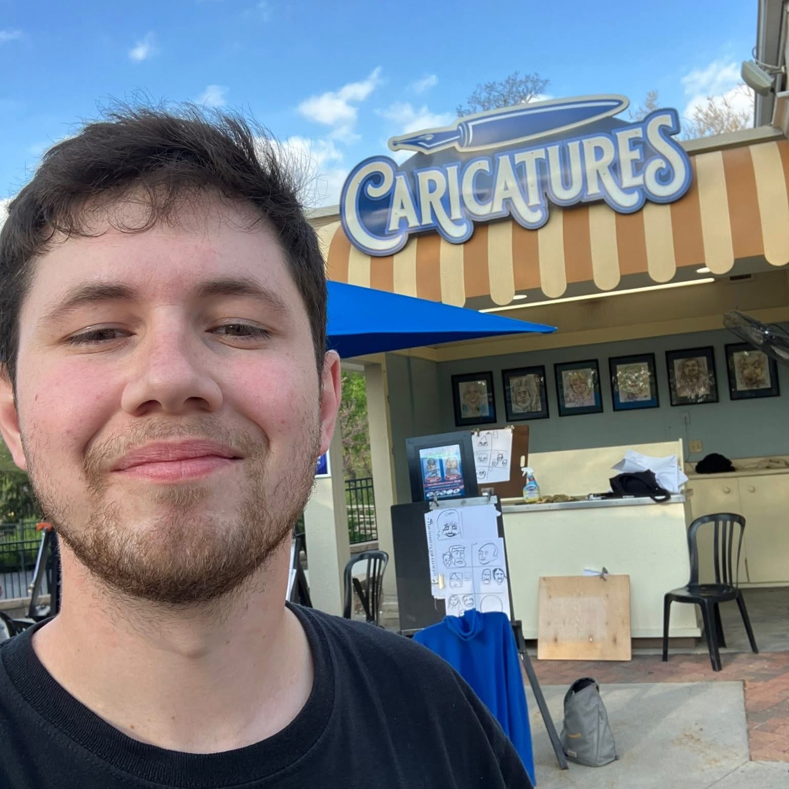 Young man with short dark hair and a beard smiling outdoors in front of a caricature stand with a striped awning and framed pictures.