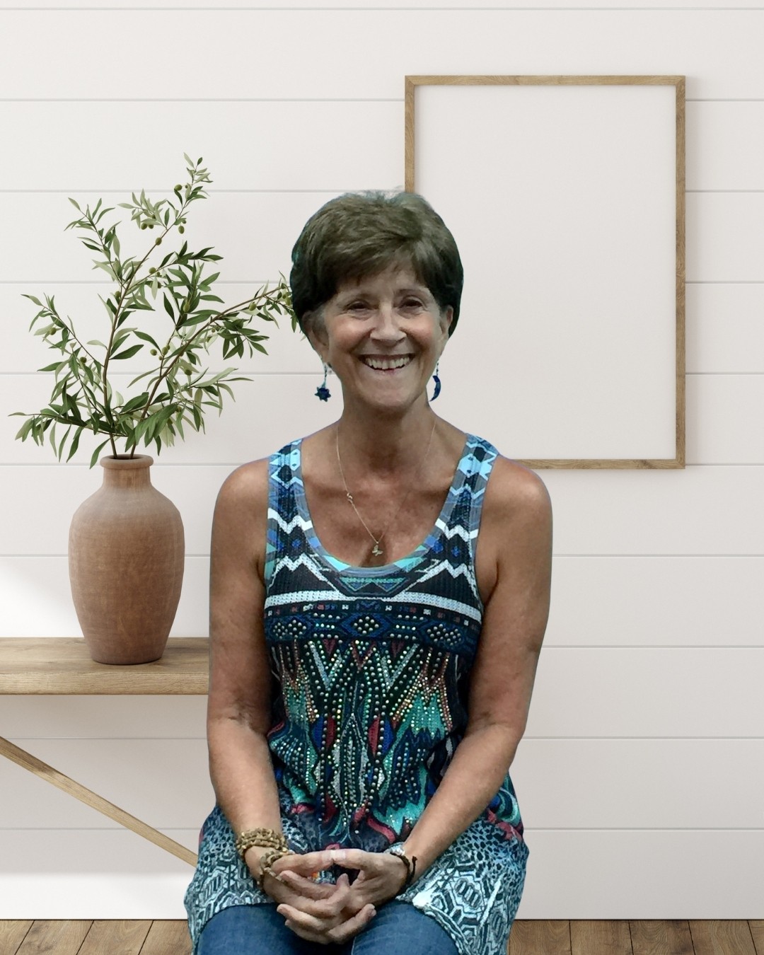 Smiling woman with short hair wearing a colorful sleeveless top, standing indoors with a potted plant and empty frame behind her.