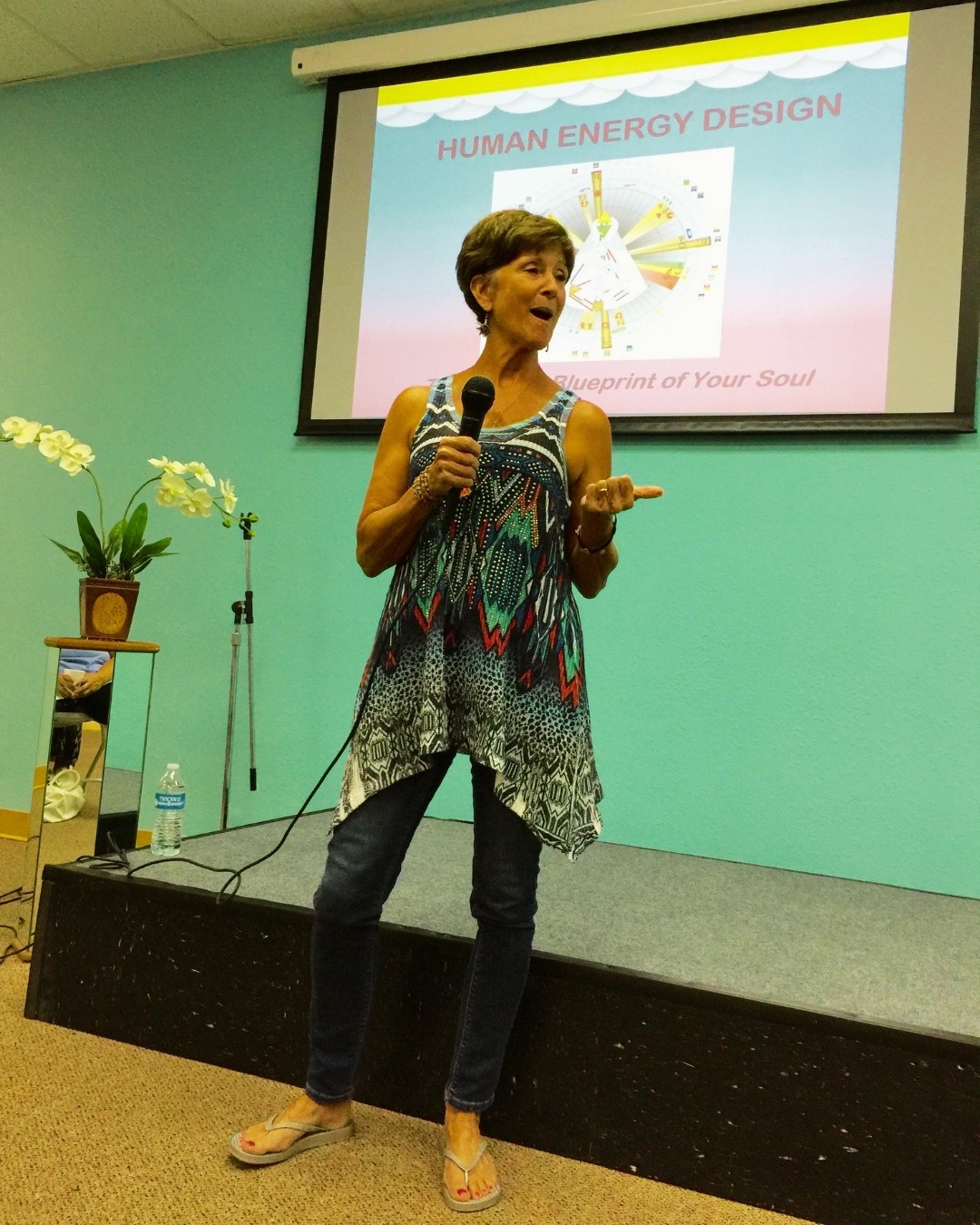Woman speaking into a microphone on stage with a presentation slide behind her, in a room with light green walls.