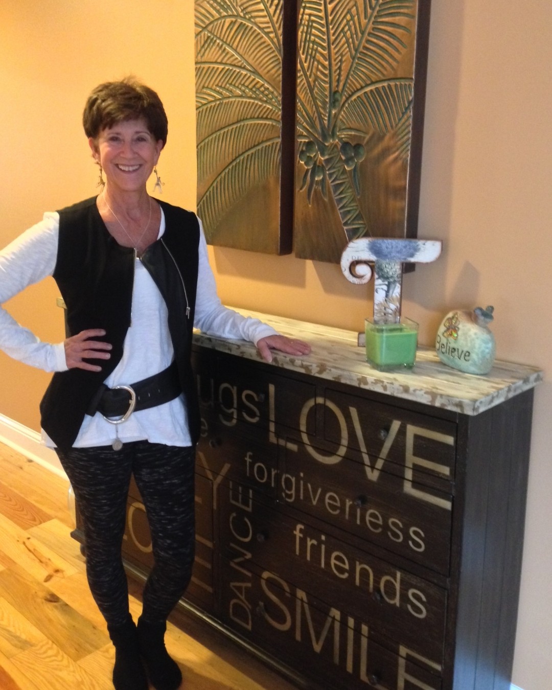 Woman standing indoors, smiling, with decorative wall art and a black cabinet with words like 'LOVE' and 'SMILE' behind her.