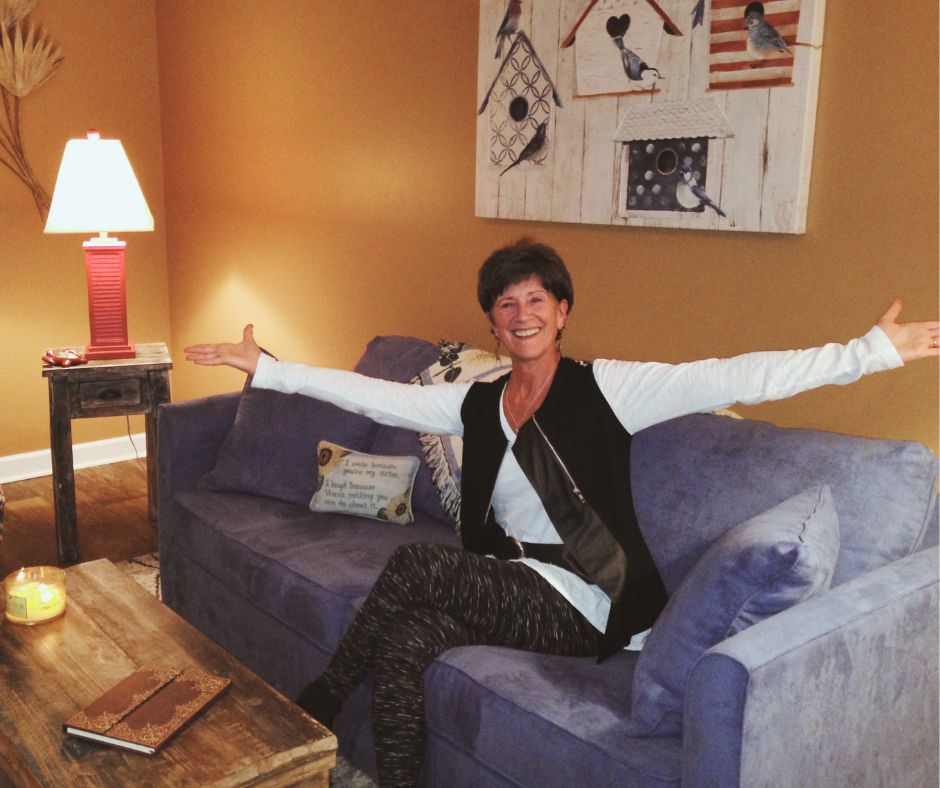 Woman sitting on a blue sofa with arms outstretched, smiling in a warmly lit living room with artwork and a lamp.