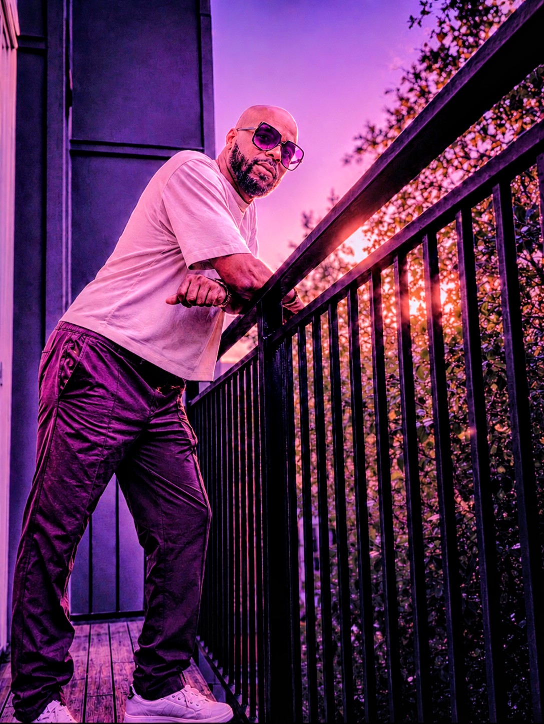 Bald man with sunglasses leaning on a railing during sunset, purple and pink sky, trees in background.