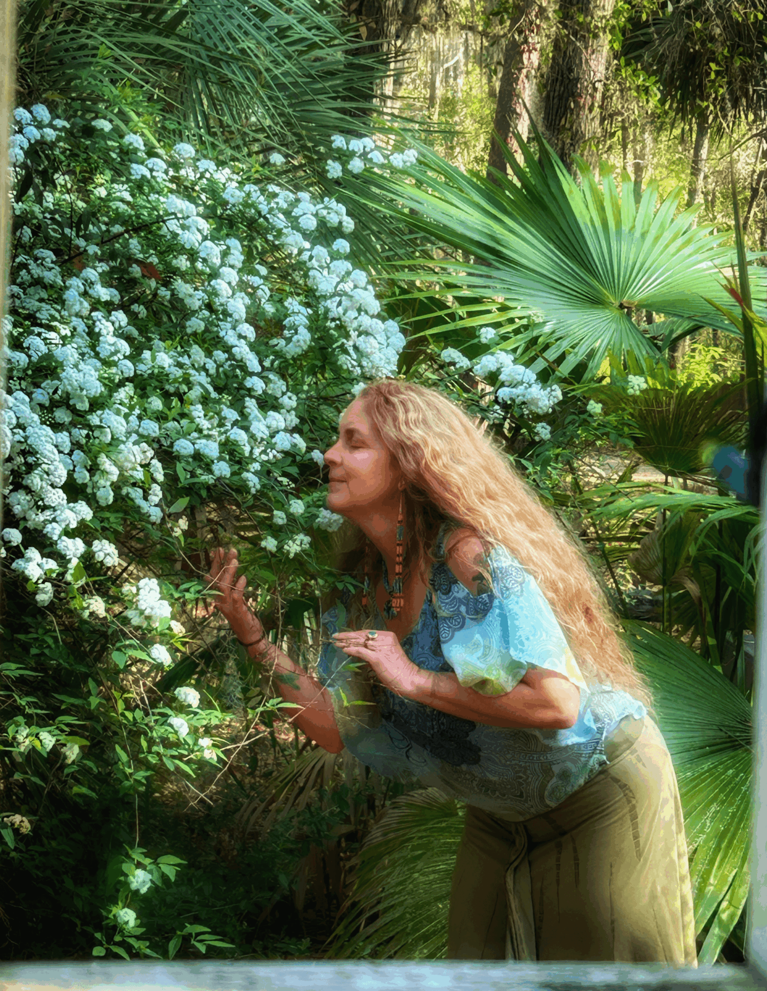 Woman with long curly hair leaning toward white flowering bush in lush green garden.