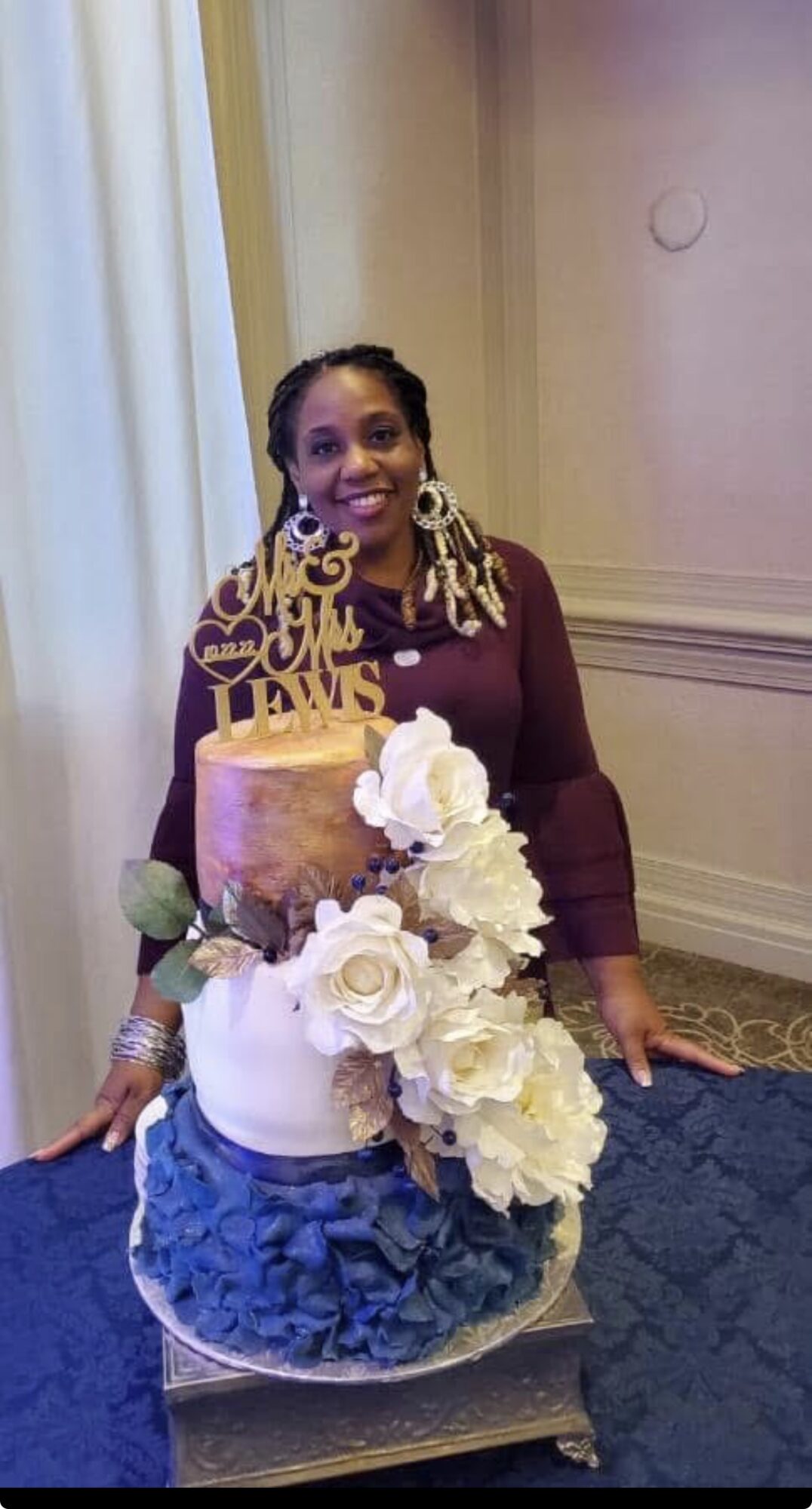Woman standing behind a three-tiered cake with white and blue flowers, smiling, in a decorated room.