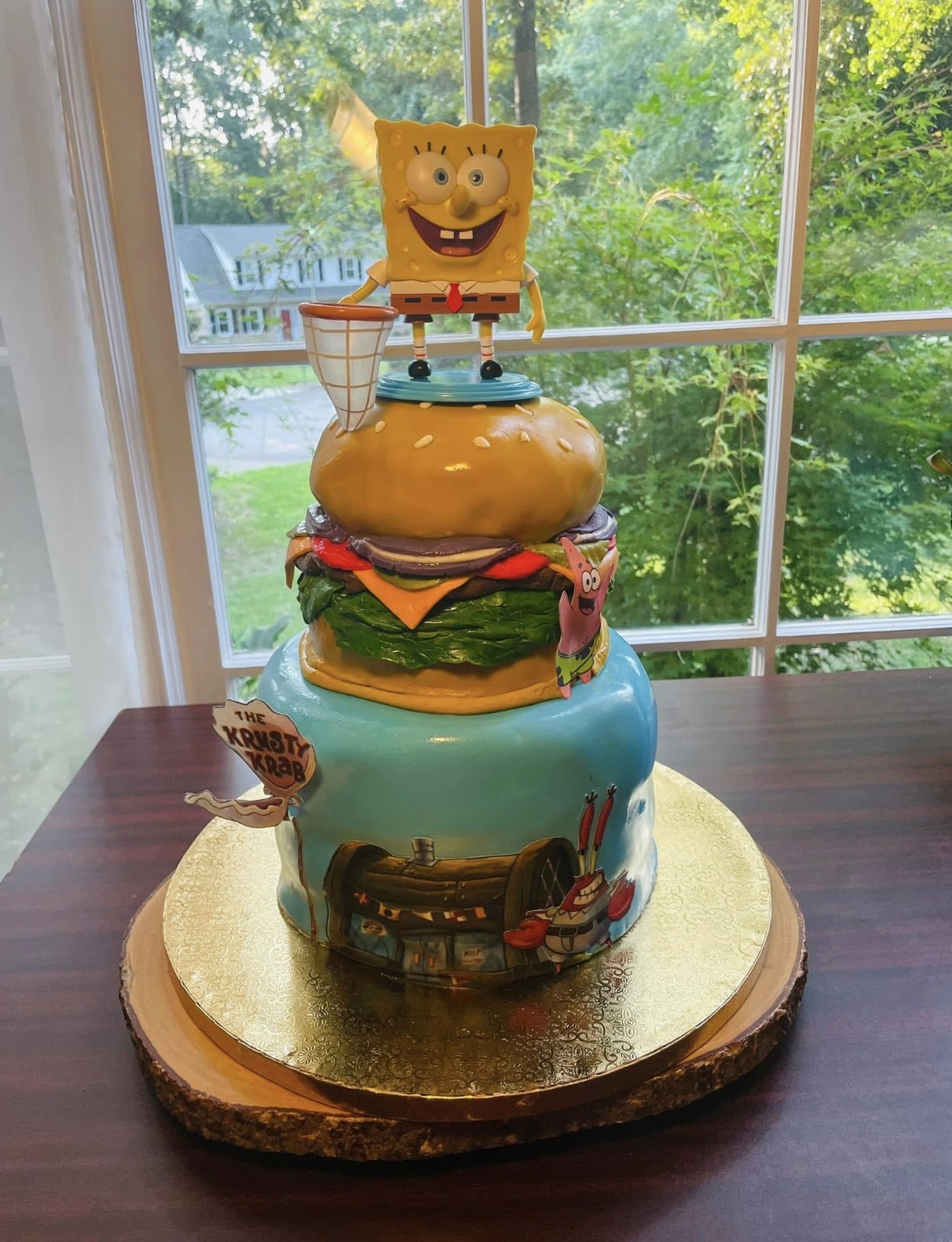Cake decorated with SpongeBob SquarePants and burger-themed elements, placed on a wooden table near a window with trees outside.