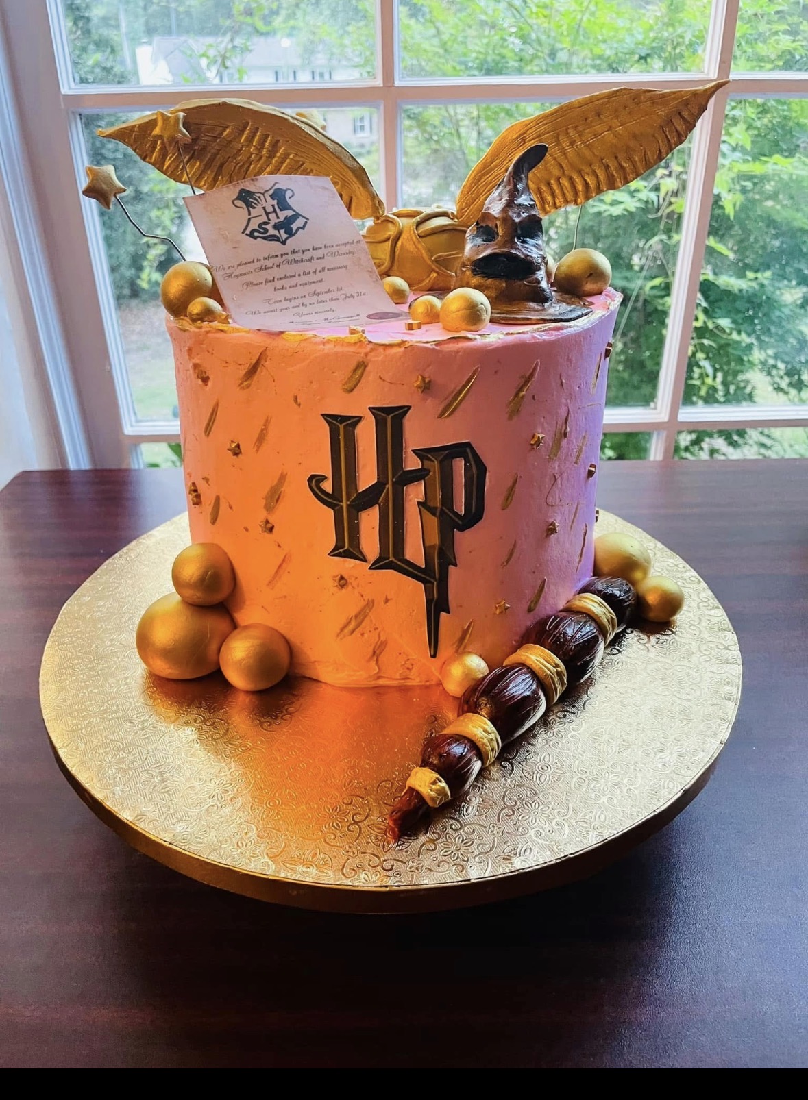 A round cake with orange and gold decorations, featuring the Harry Potter logo, two golden wings, and a scroll on top.