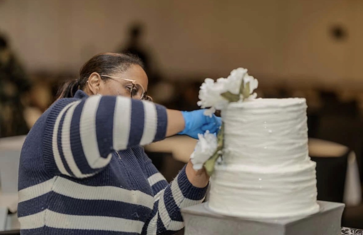 Person decorating a large white tiered cake with flowers, wearing glasses, a striped sweater, and blue gloves.