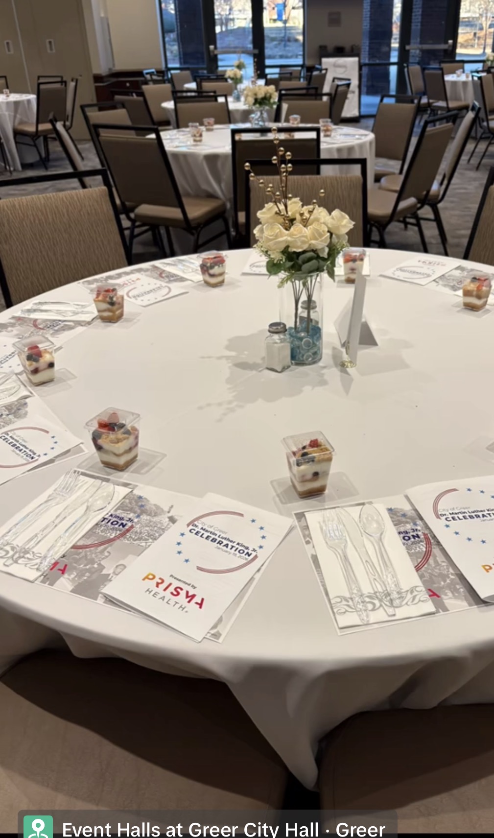 Round table with white tablecloth, floral centerpiece, cupcakes, and event materials, in a decorated event hall.