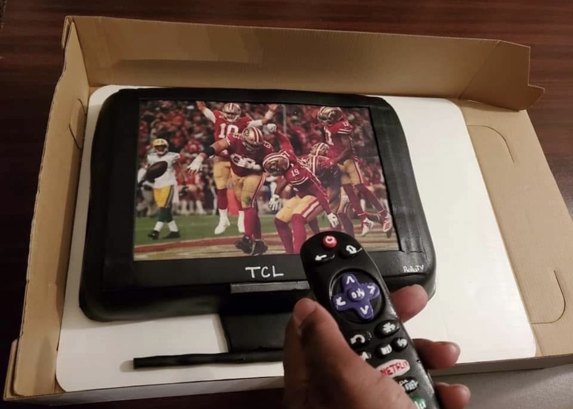 Portable TV displaying a football game, held by a person with a remote control, inside a cardboard box.