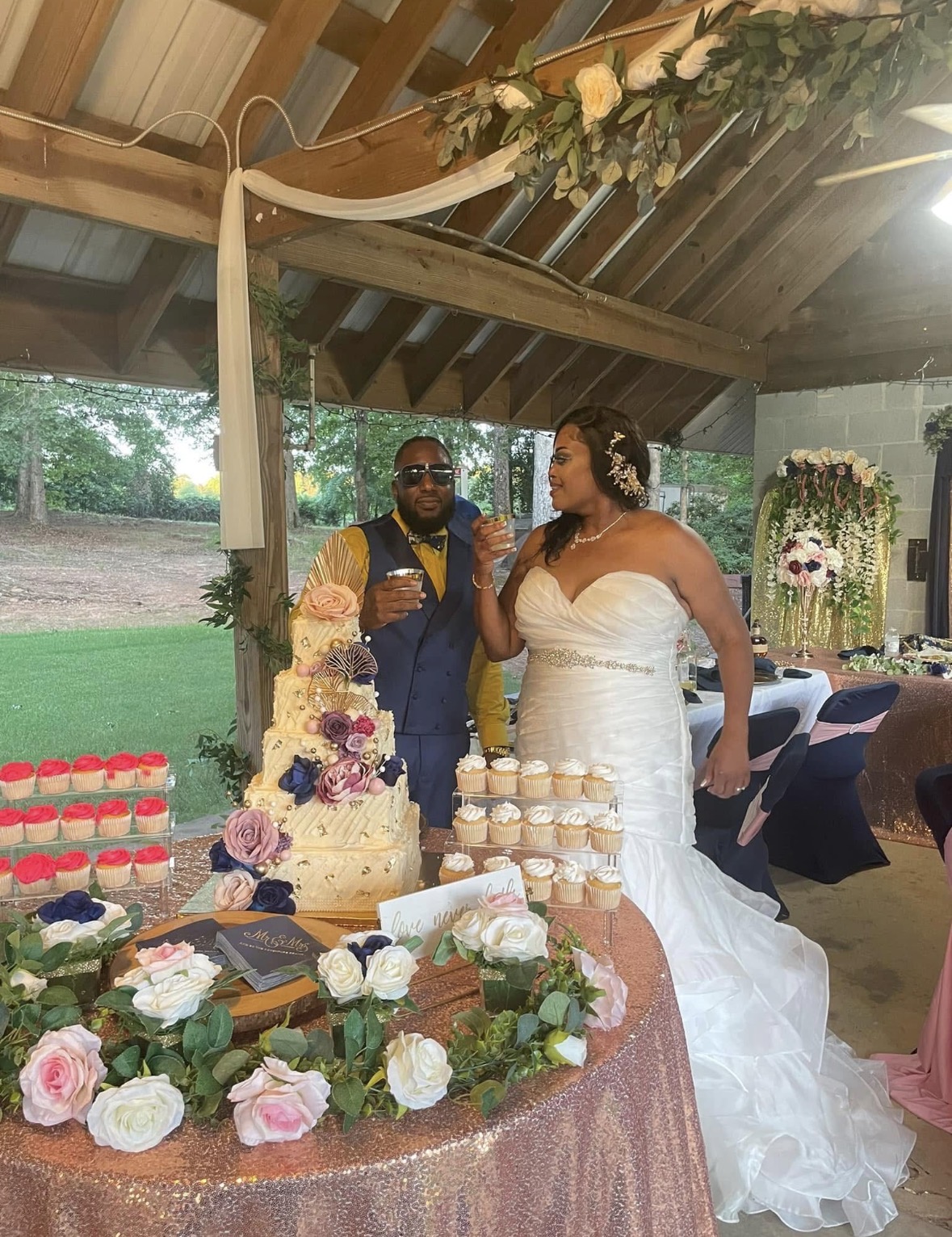 Bride and groom stand near wedding cake under decorated wooden pavilion, surrounded by flowers and wedding decorations.
