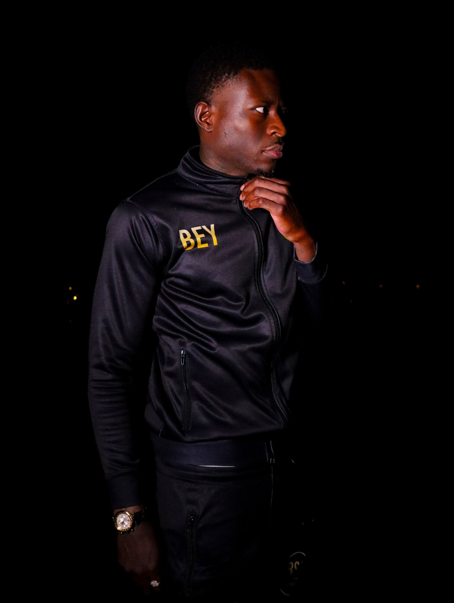 Young man in black jacket with yellow text, standing sideways with hand near chin, against black background.