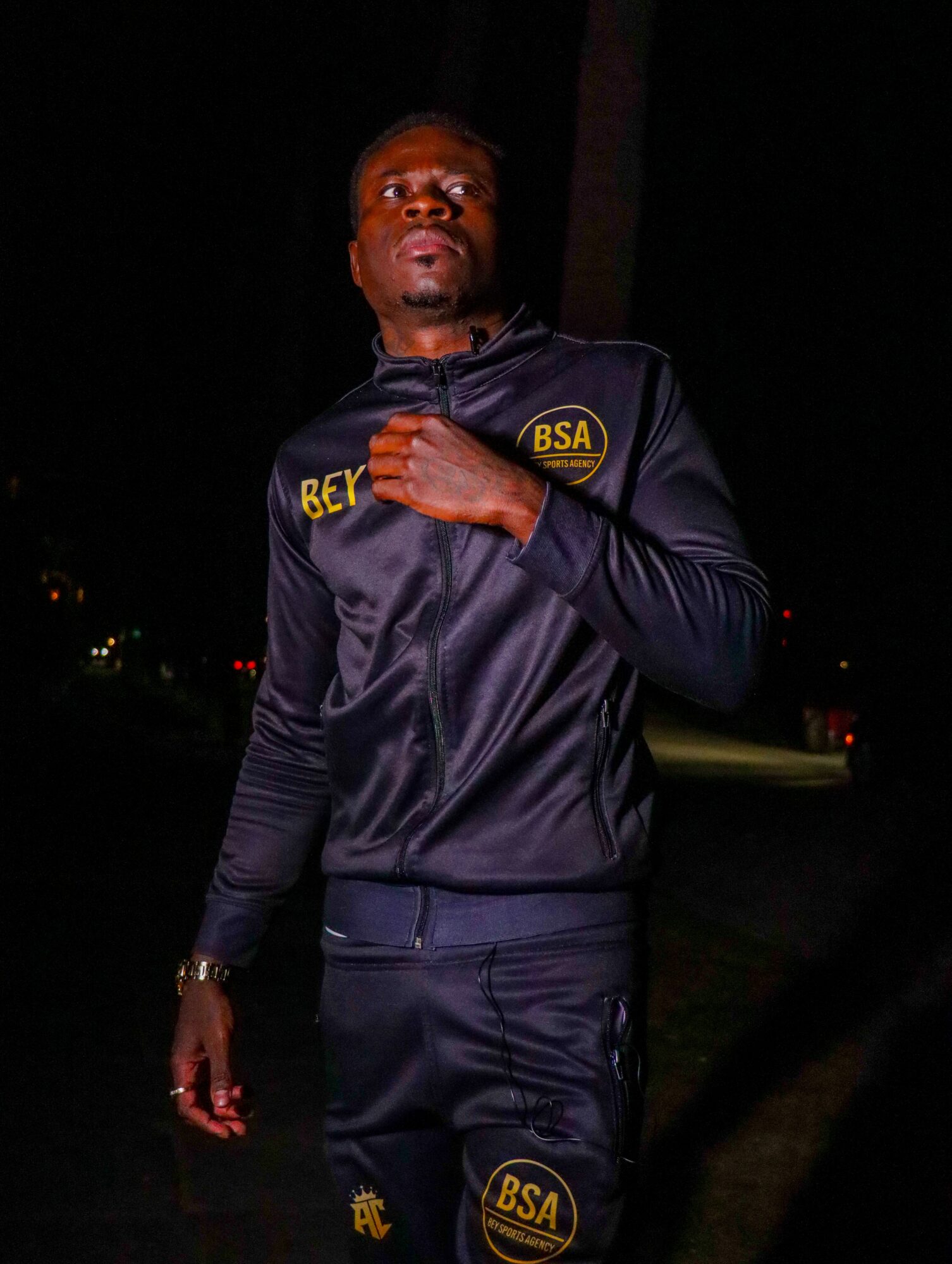 Man in dark sports jacket standing outdoors at night, hand on chest, looking at camera.