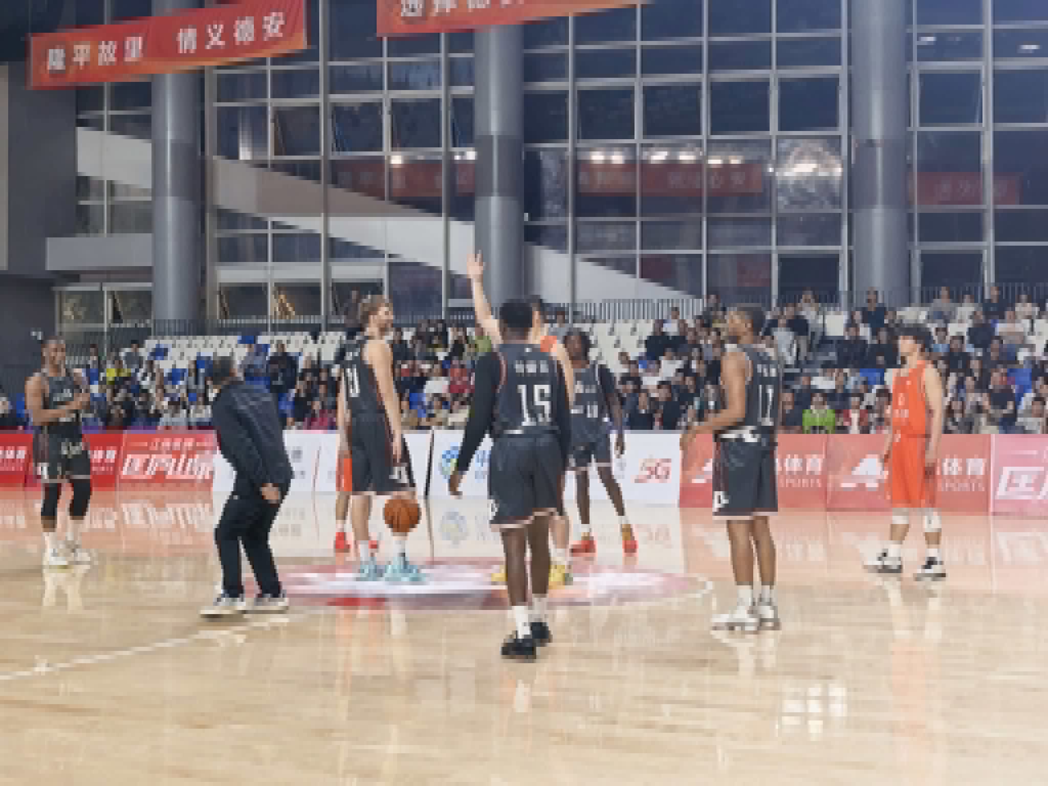 Basketball players on court with spectators in background, some players in black jerseys, some in orange, coach nearby.