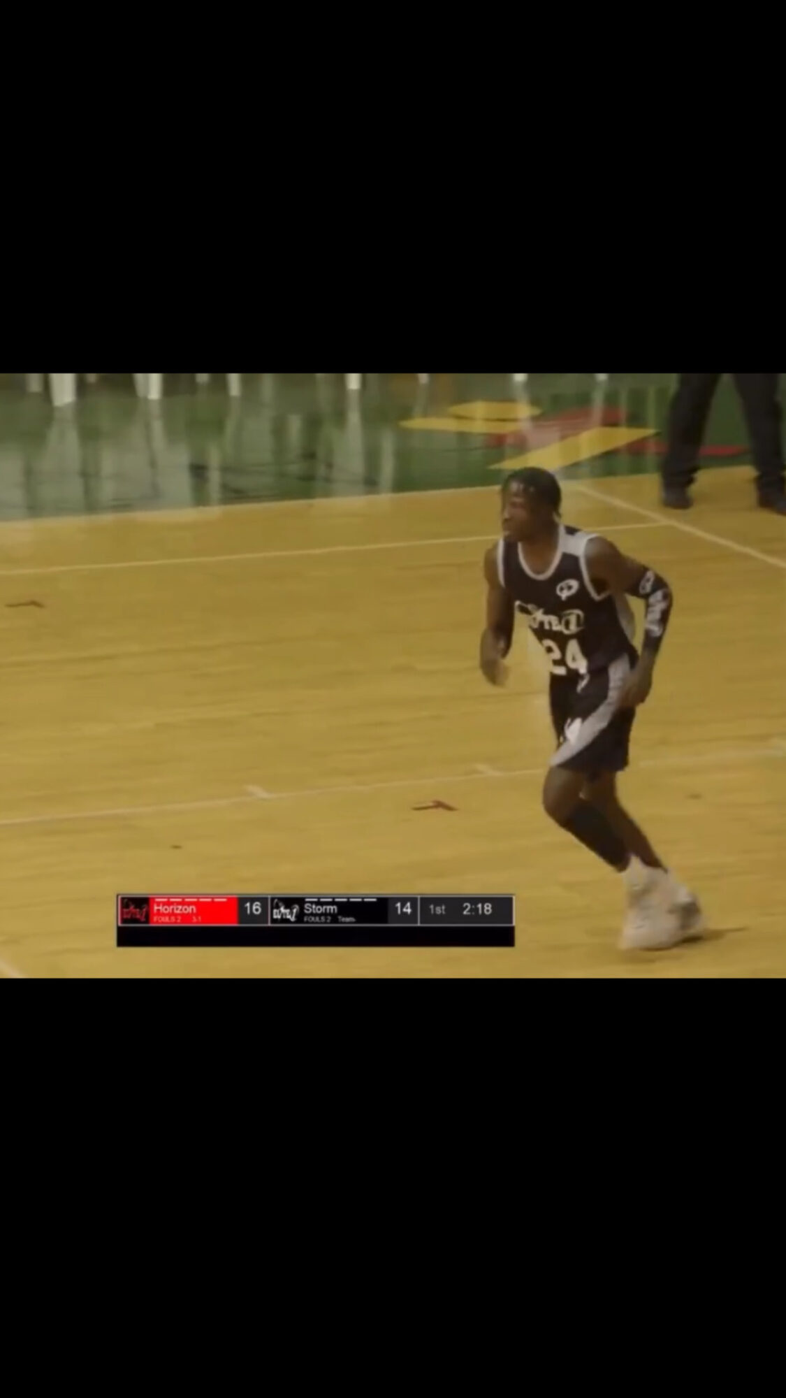 Basketball player in uniform dribbling on court during game, with scoreboard visible below, background shows court markings.