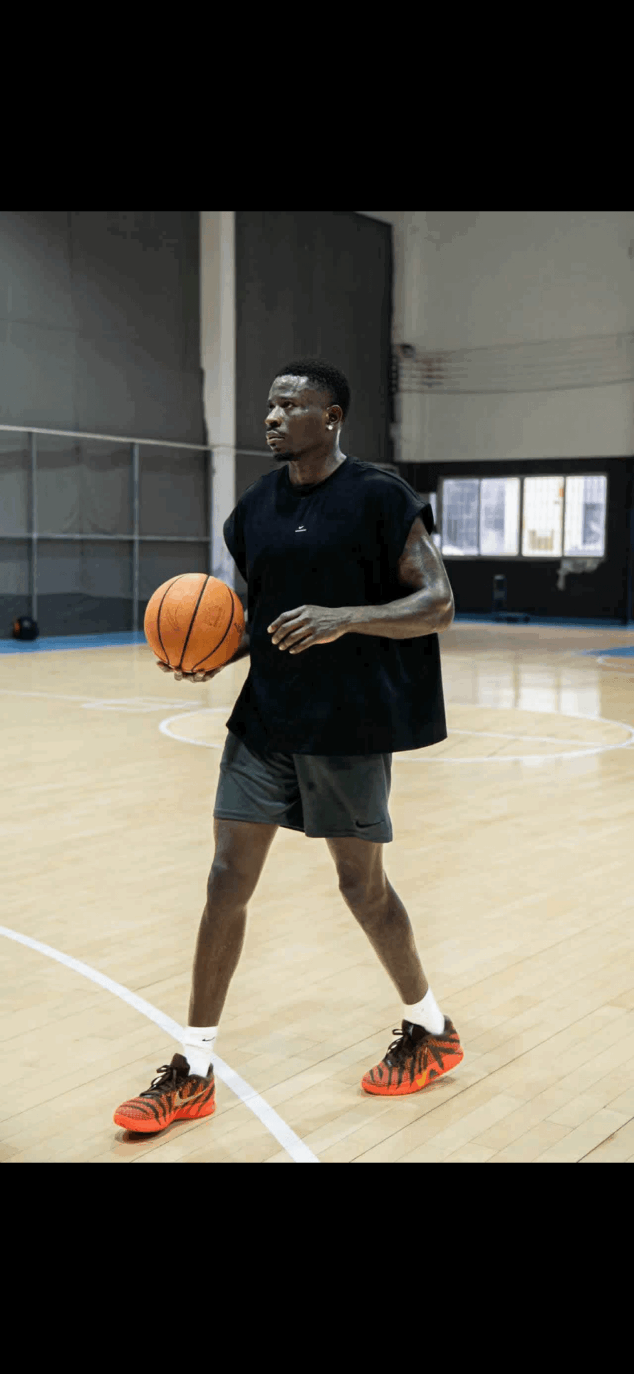 Person holding basketball on indoor court, wearing black shirt, green shorts, and orange shoes, smiling, with windows in background.