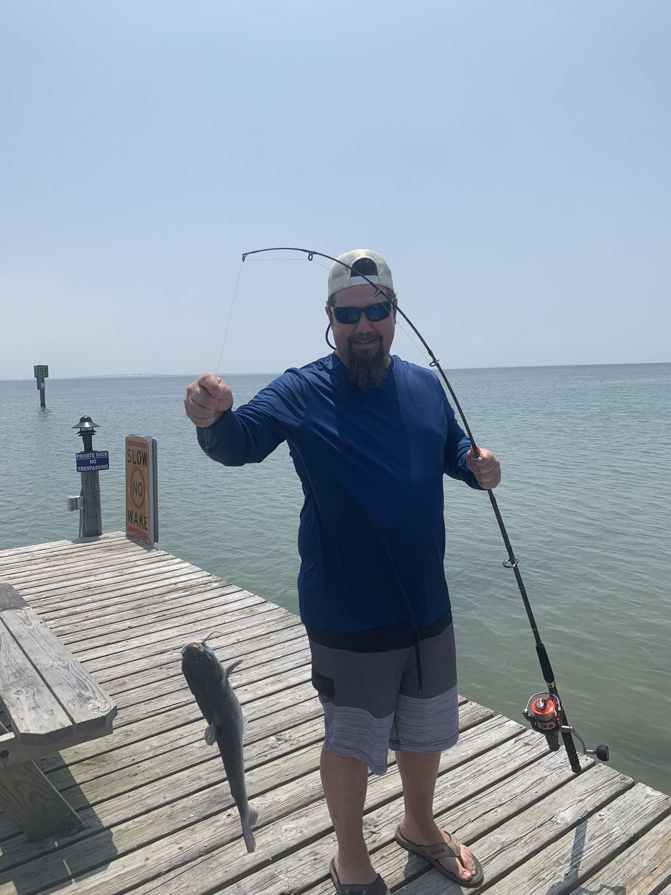Man standing on a wooden dock holding a fishing rod with a fish hanging from the line, water and sky in background.