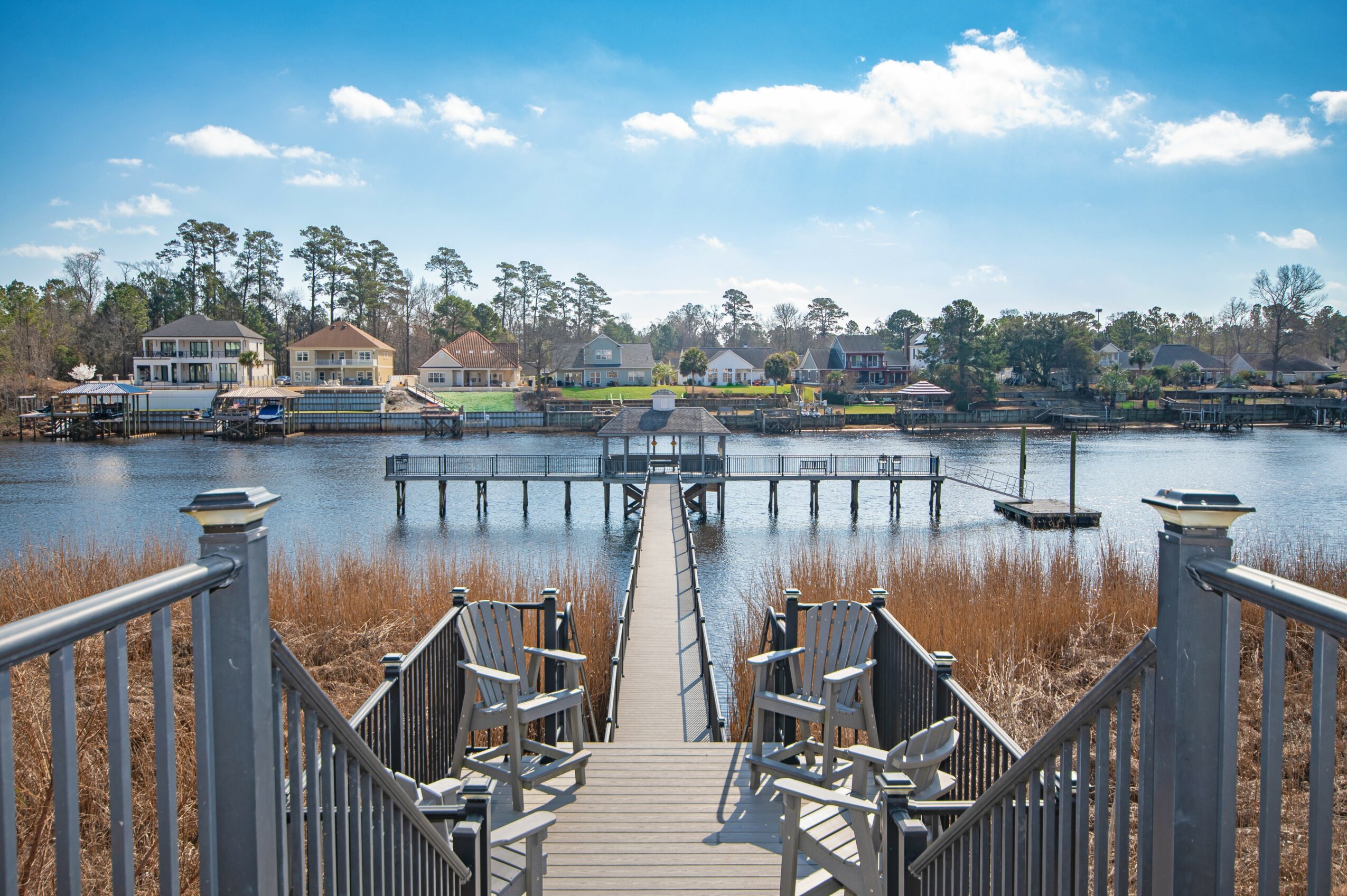 View of a dock leading to a waterway with houses and trees in the background, under a partly cloudy sky.