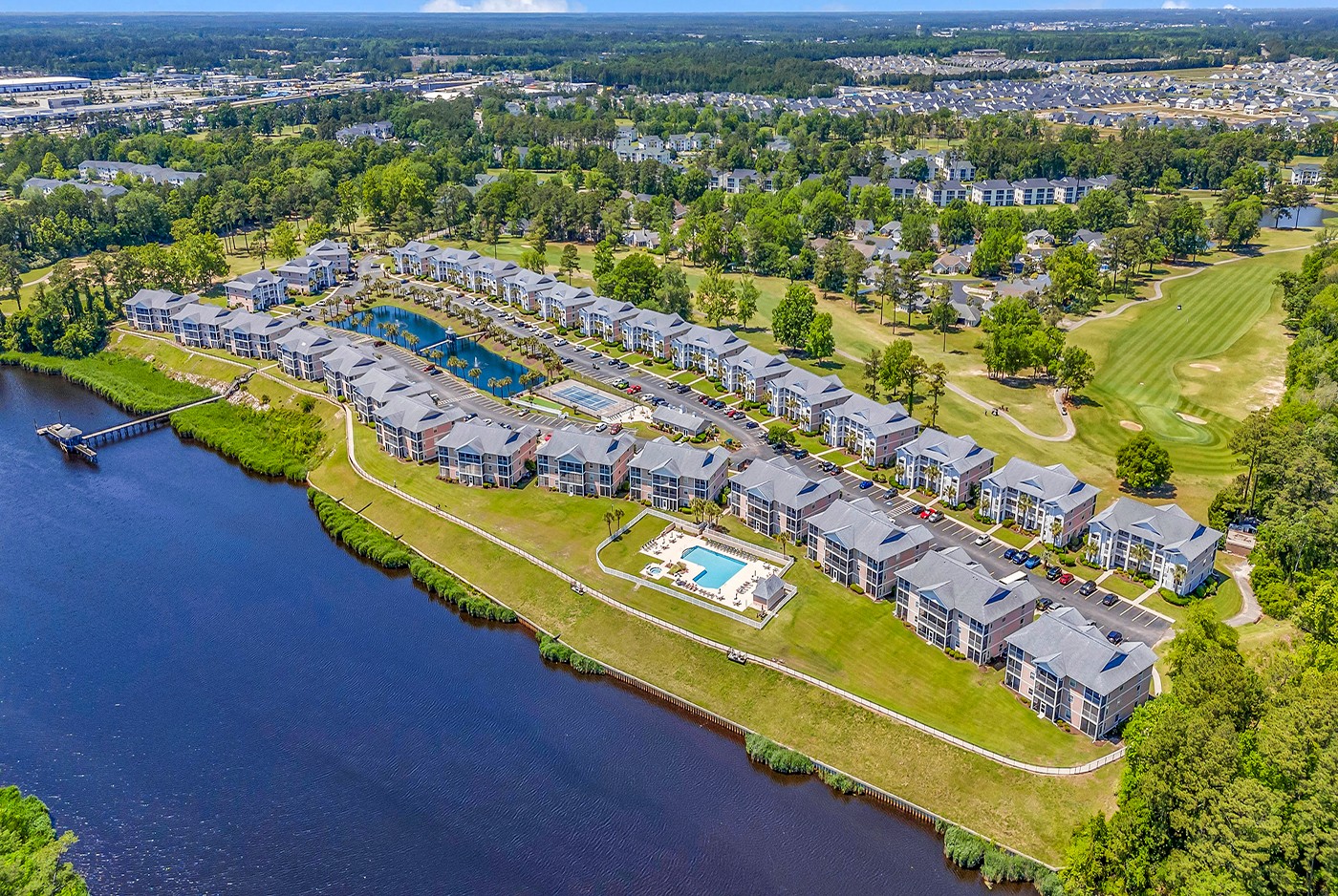 Aerial view of a residential community with townhouses, a swimming pool, and a lake, surrounded by greenery and open spaces.