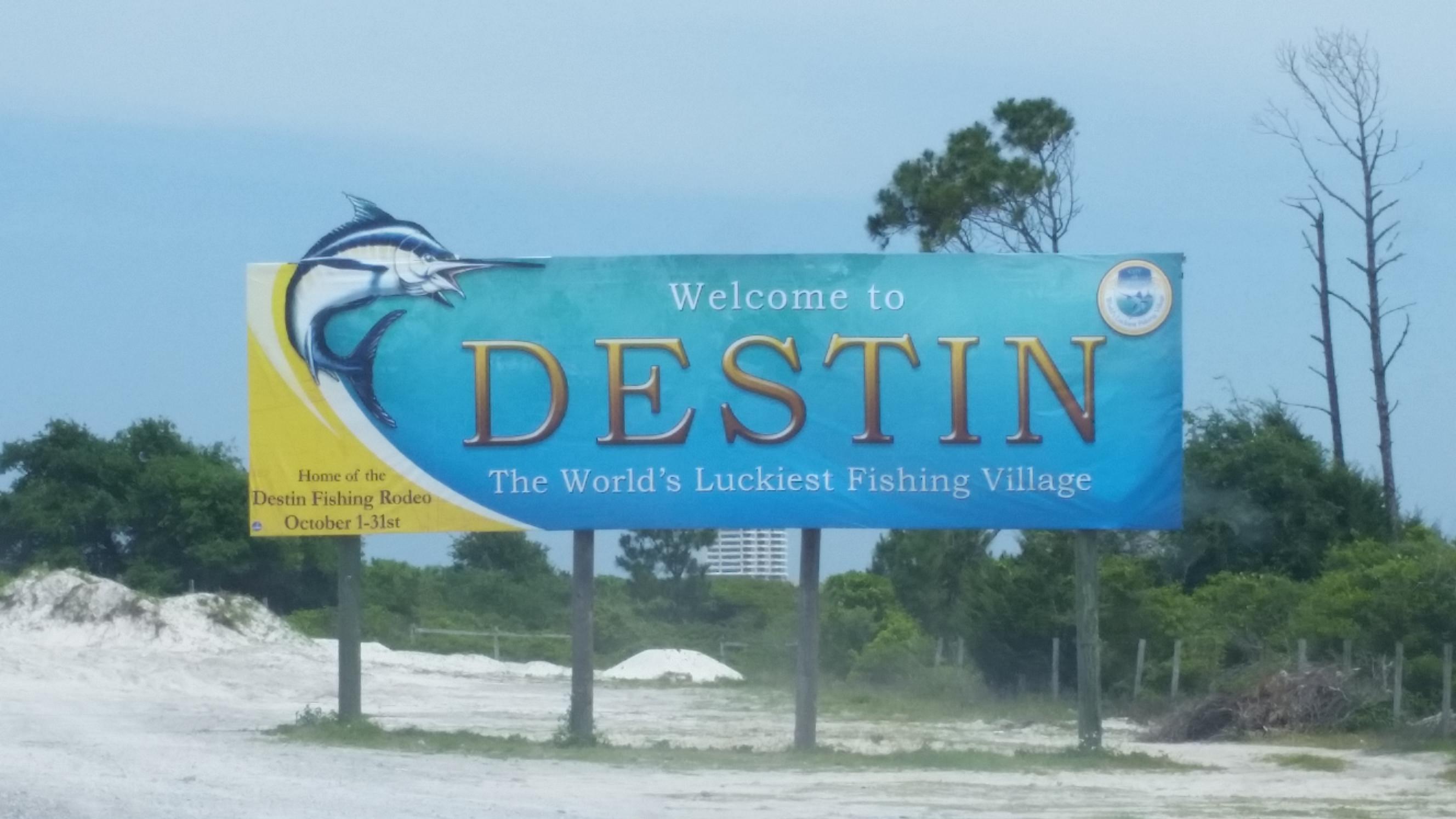 Welcome to DESTIN sign with a fish graphic, trees, and sky in the background.