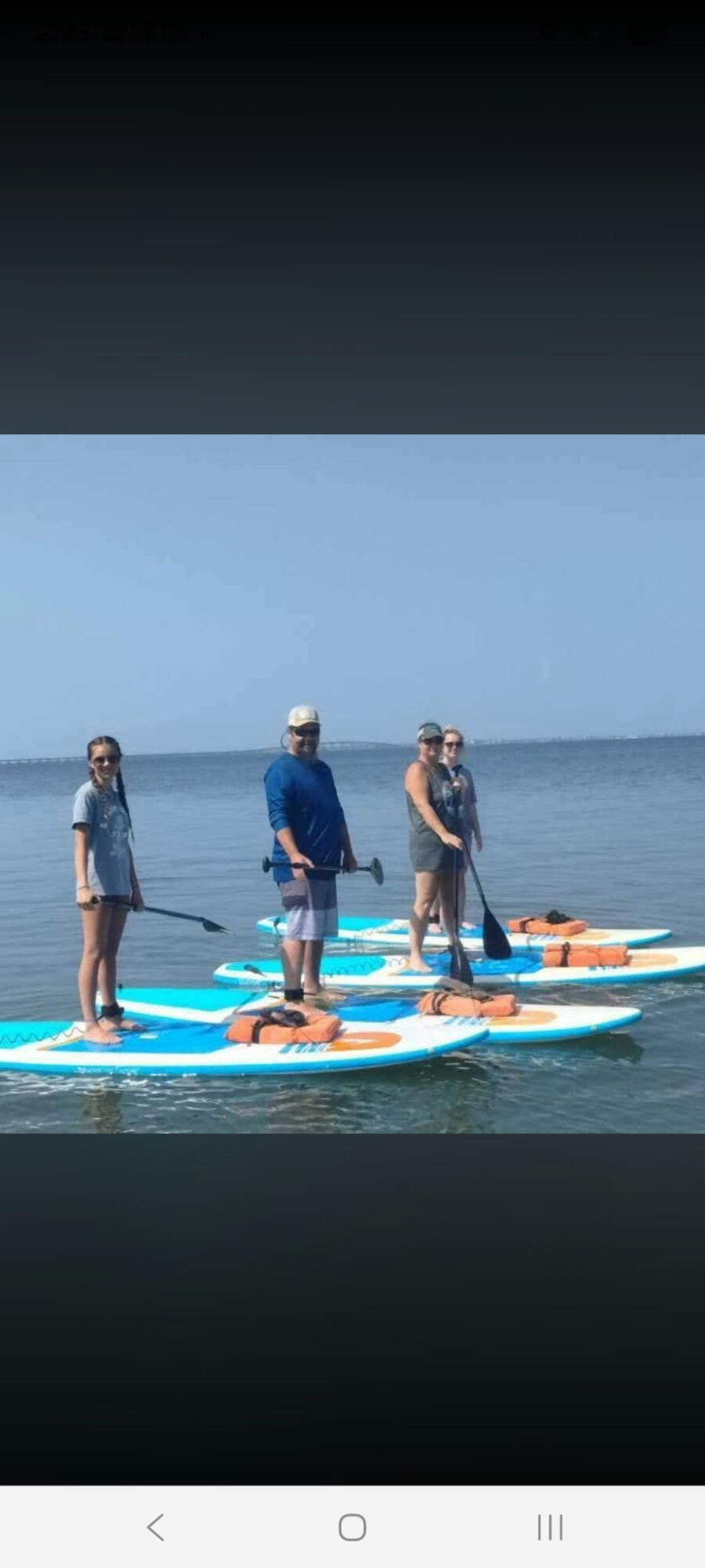 Four people stand on paddleboards in the water, facing the camera, with a clear sky and ocean background.