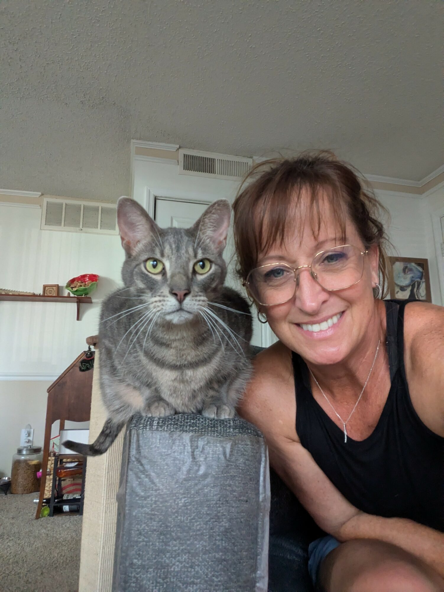 Smiling woman with glasses next to a gray tabby cat on a ledge in a home interior.