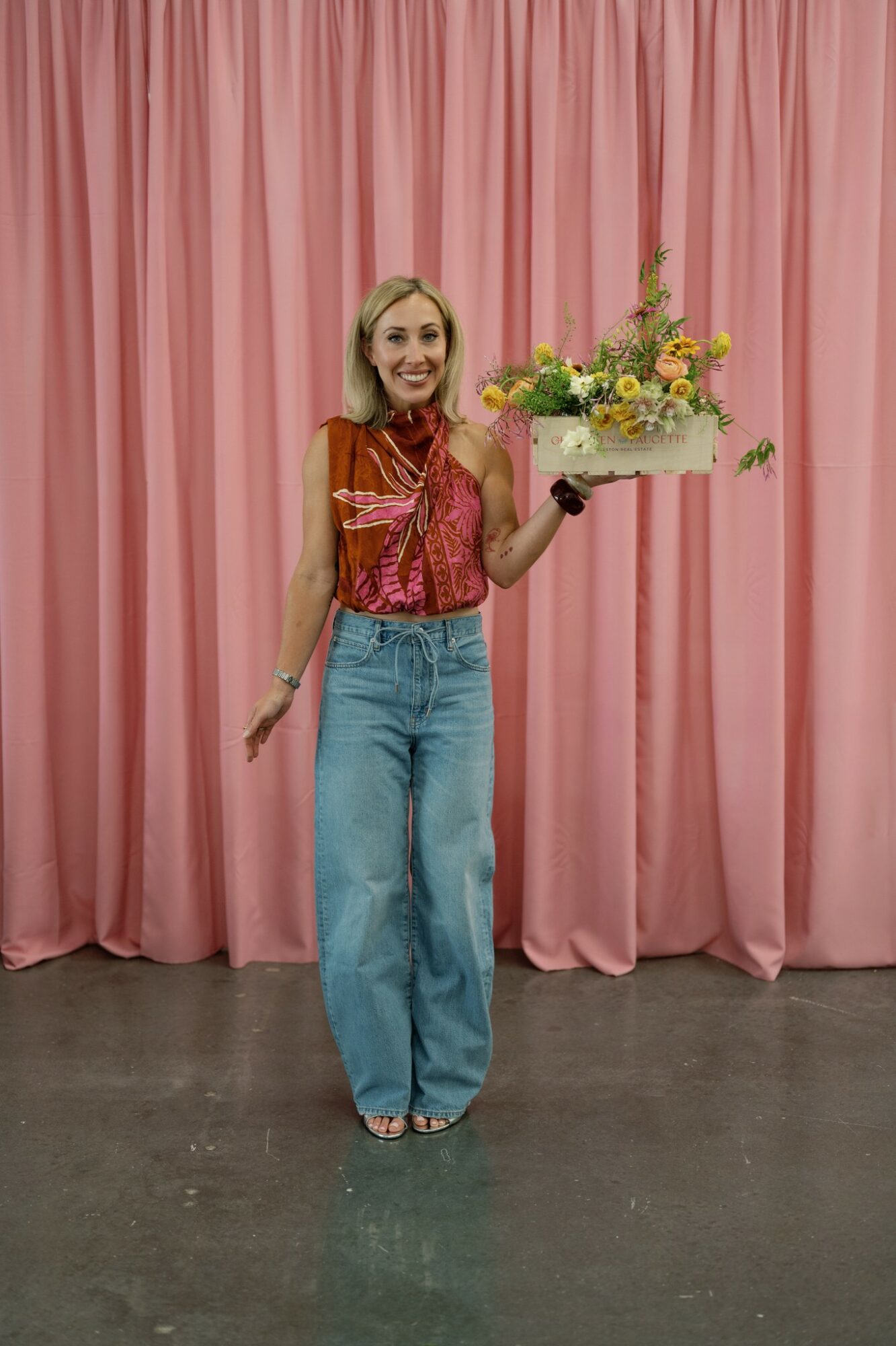 Woman holding a large flower arrangement in front of pink curtains, smiling, wearing a red top and blue jeans.